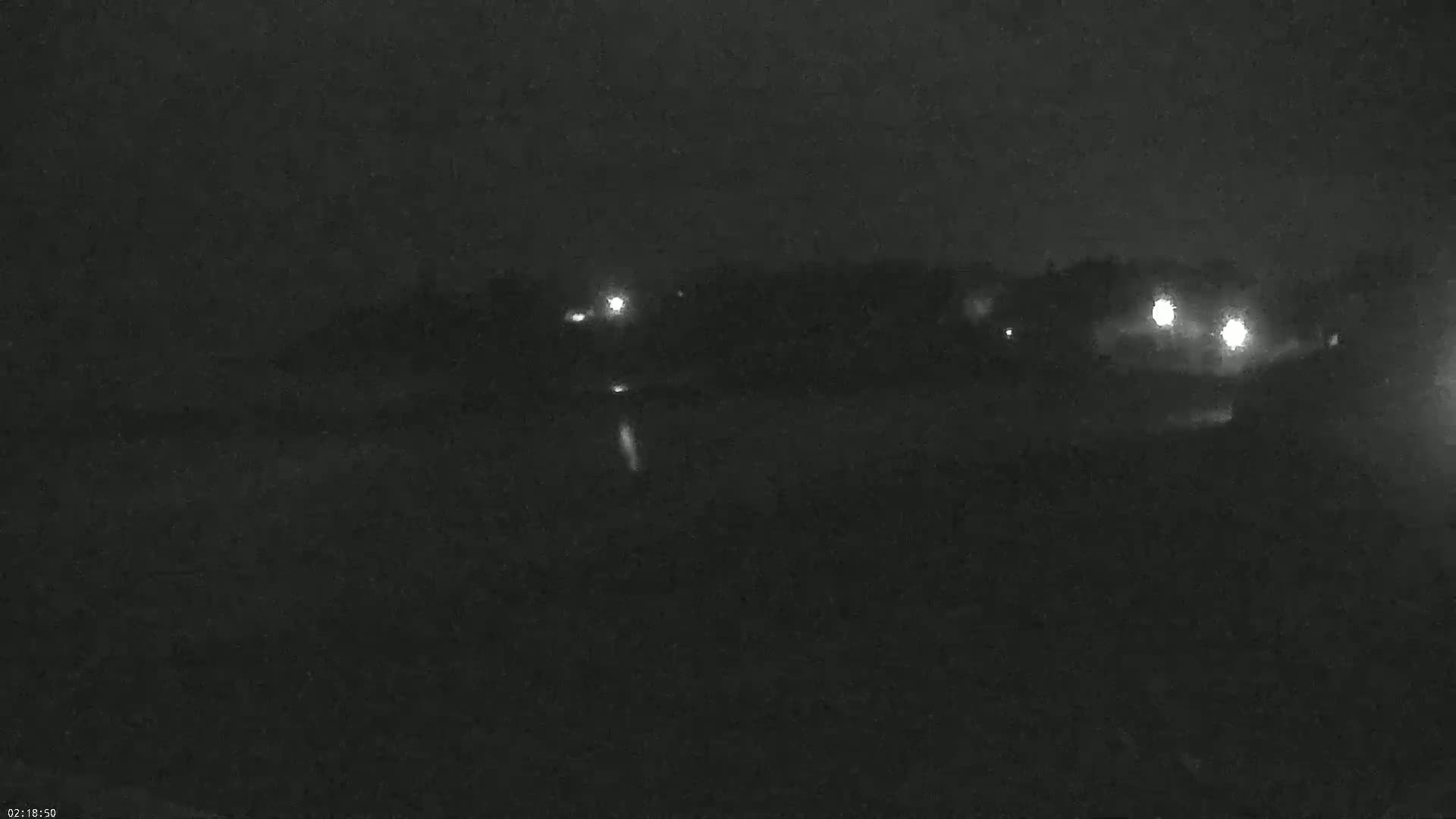 A dark, nighttime scene shows a few distant lights behind a dark body of water under a cloudy sky.