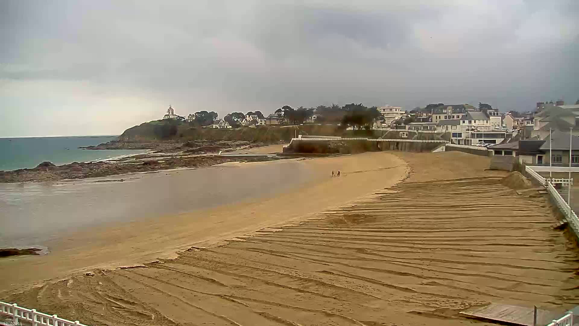A sandy beach with tire tracks stretches toward a calm bay and a coastal town under an overcast sky.