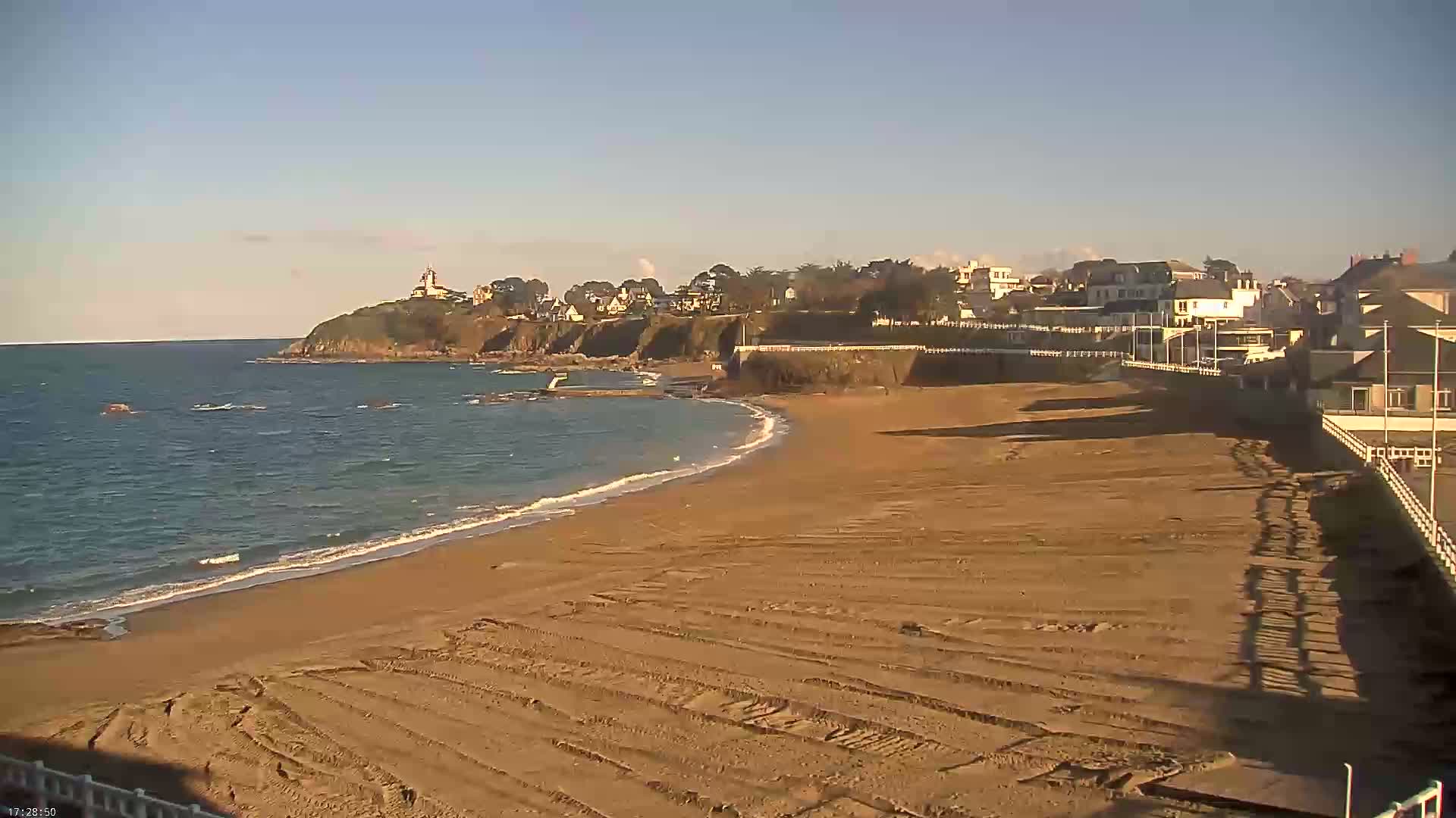 A sandy beach stretches along a calm ocean under a clear sky, with houses and a small cliff visible in the background.