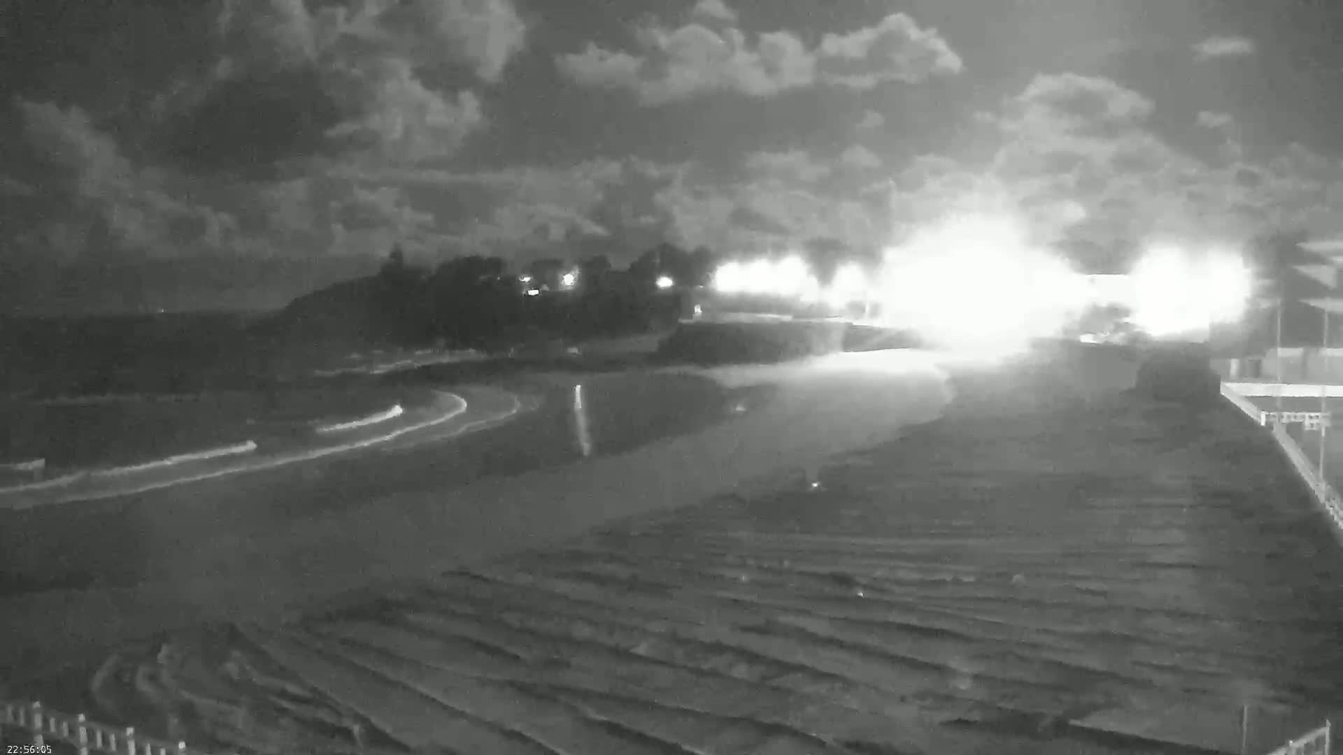 A nighttime, overcast scene shows a paved roadway curving through a dark landscape, illuminated by bright lights in the distance.
