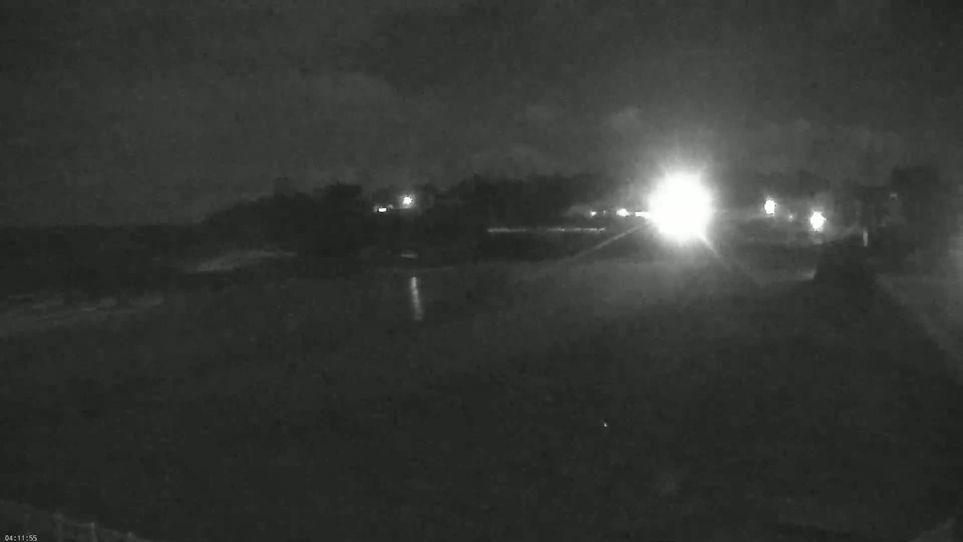 A nighttime, low-light view shows a road with headlights from vehicles in the distance, and some dimly lit buildings or trees in the background.