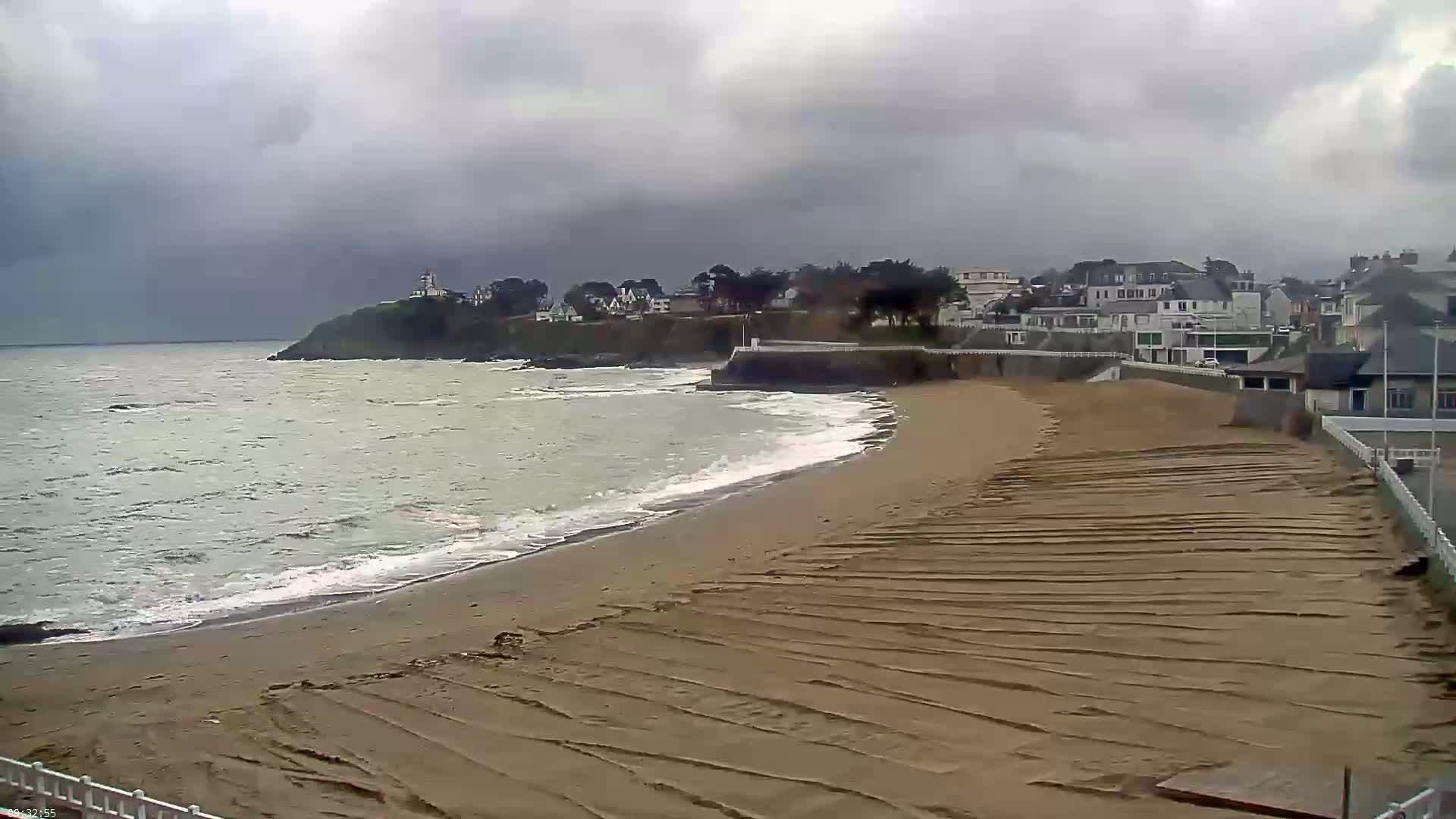 A sandy beach with textured patterns from receding waves stretches towards a shoreline with houses and a lighthouse under a cloudy sky.