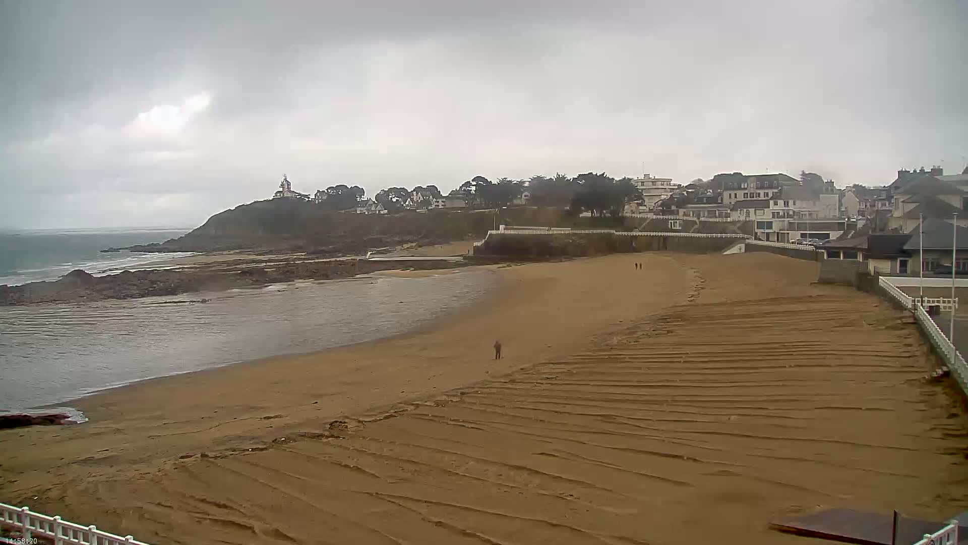 A sandy beach stretches toward a small town under an overcast sky, with a rocky shoreline and ocean visible in the background.