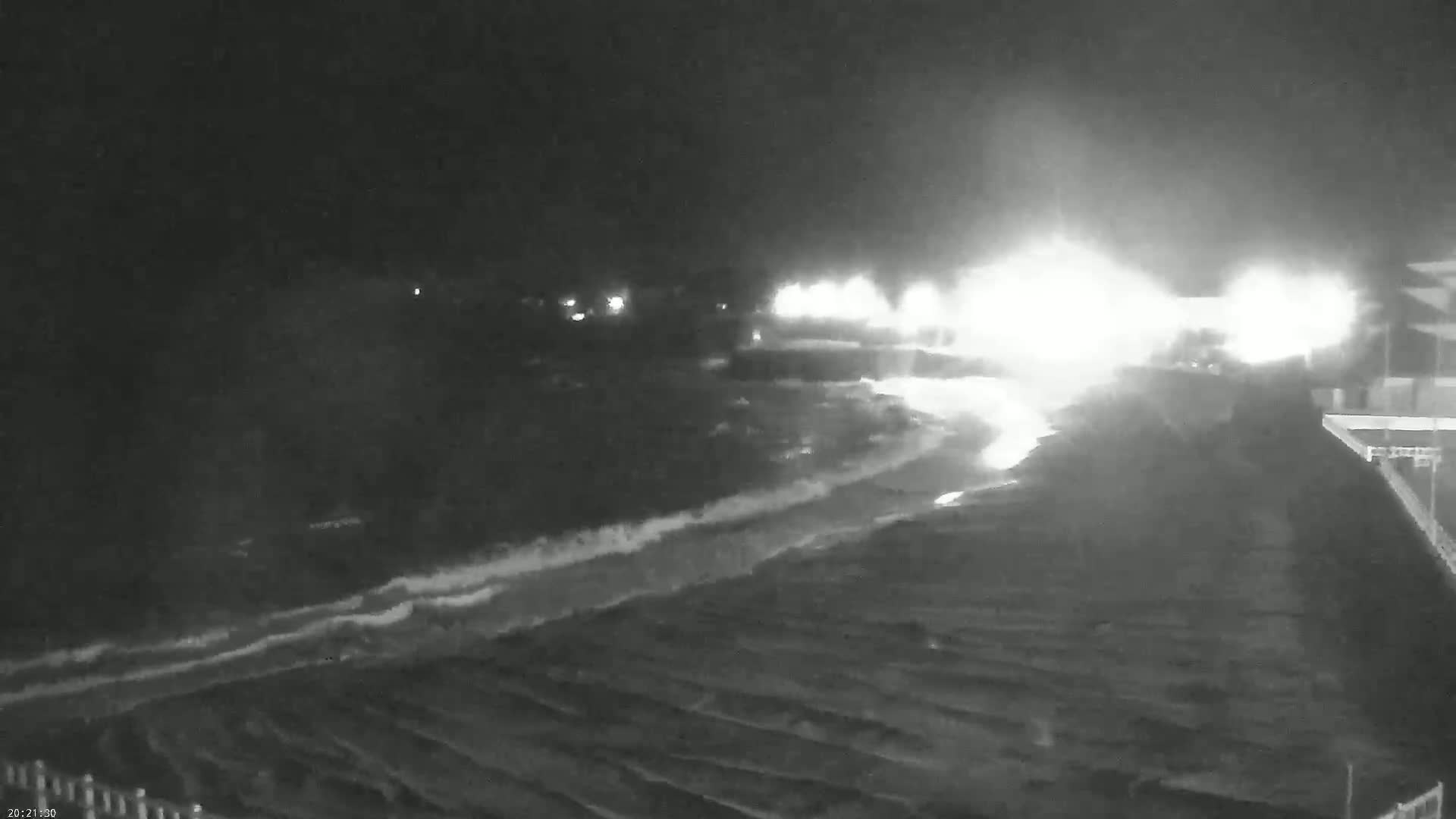 A nighttime, low-light image shows a body of water with waves crashing on a shore, with bright lights visible in the distance.