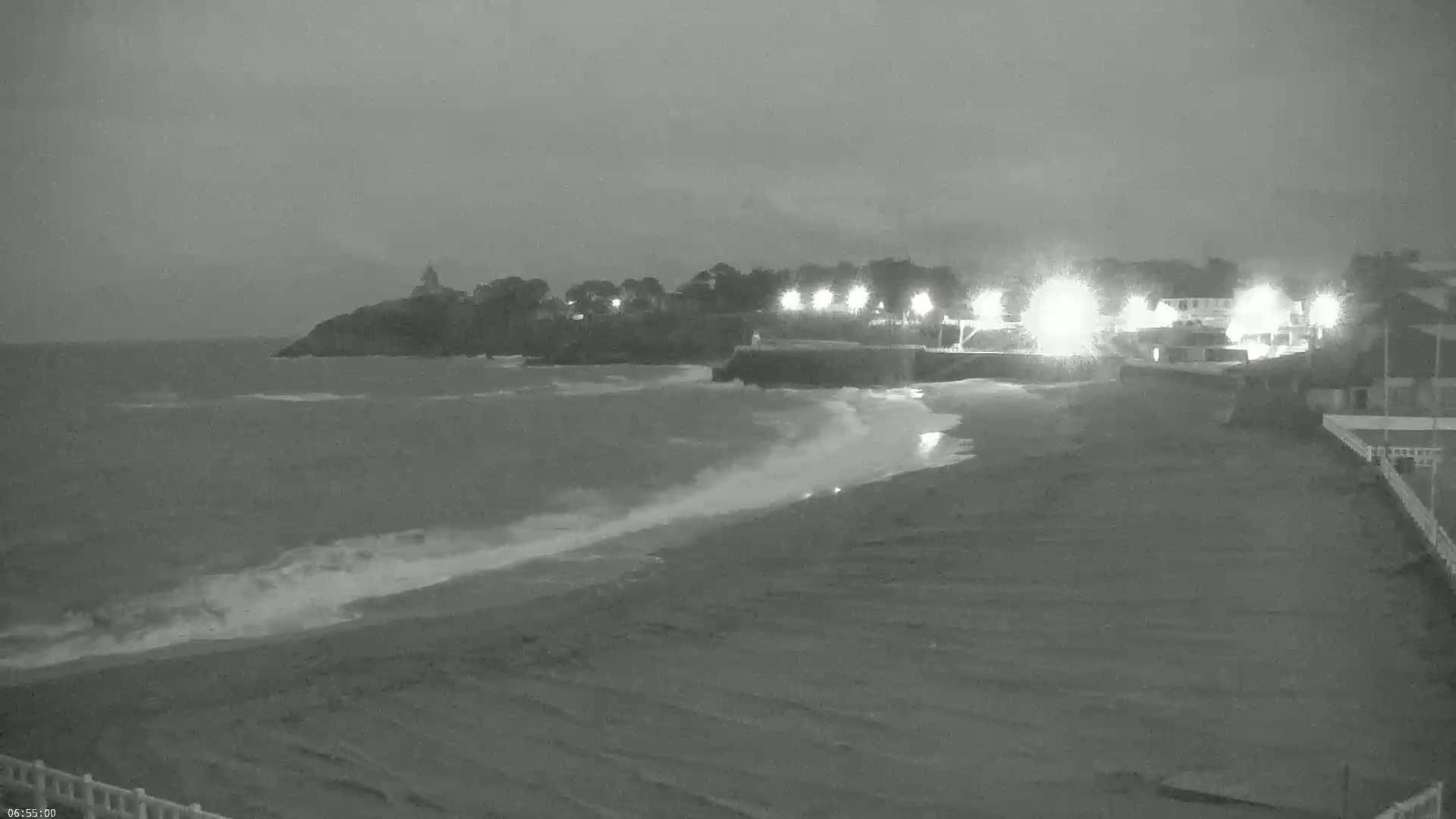Under a dark, overcast sky, small waves lap a sandy beach at night, illuminated by lights along the shore near a rocky point.