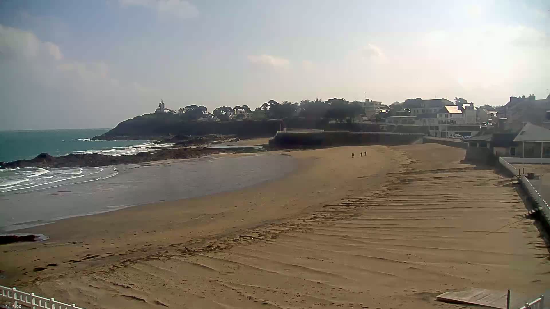 A sandy beach with gentle waves laps the shore under a partly cloudy sky, with a small town and a lighthouse visible in the distance.