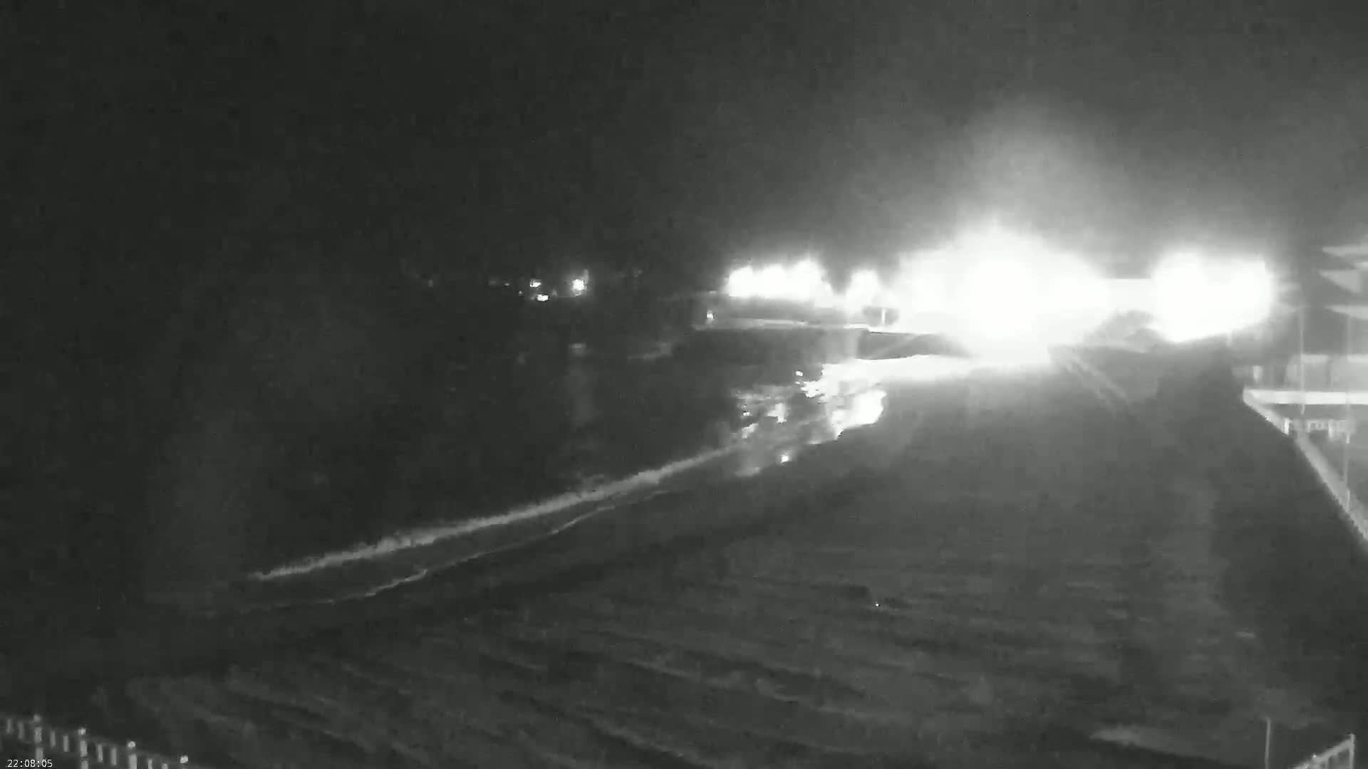 A nighttime, low-light image shows a wet, dark roadway leading to brightly lit buildings in the distance, possibly under conditions of rain or fog.