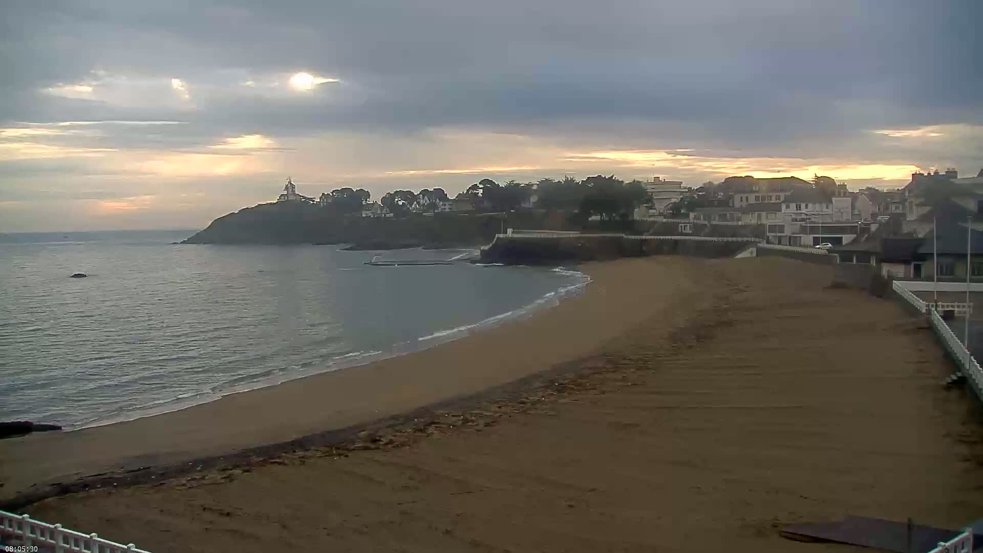 A sandy beach curves around a calm bay under a partly cloudy, sunrise sky, with a small town and a lighthouse visible on a cliff in the background.