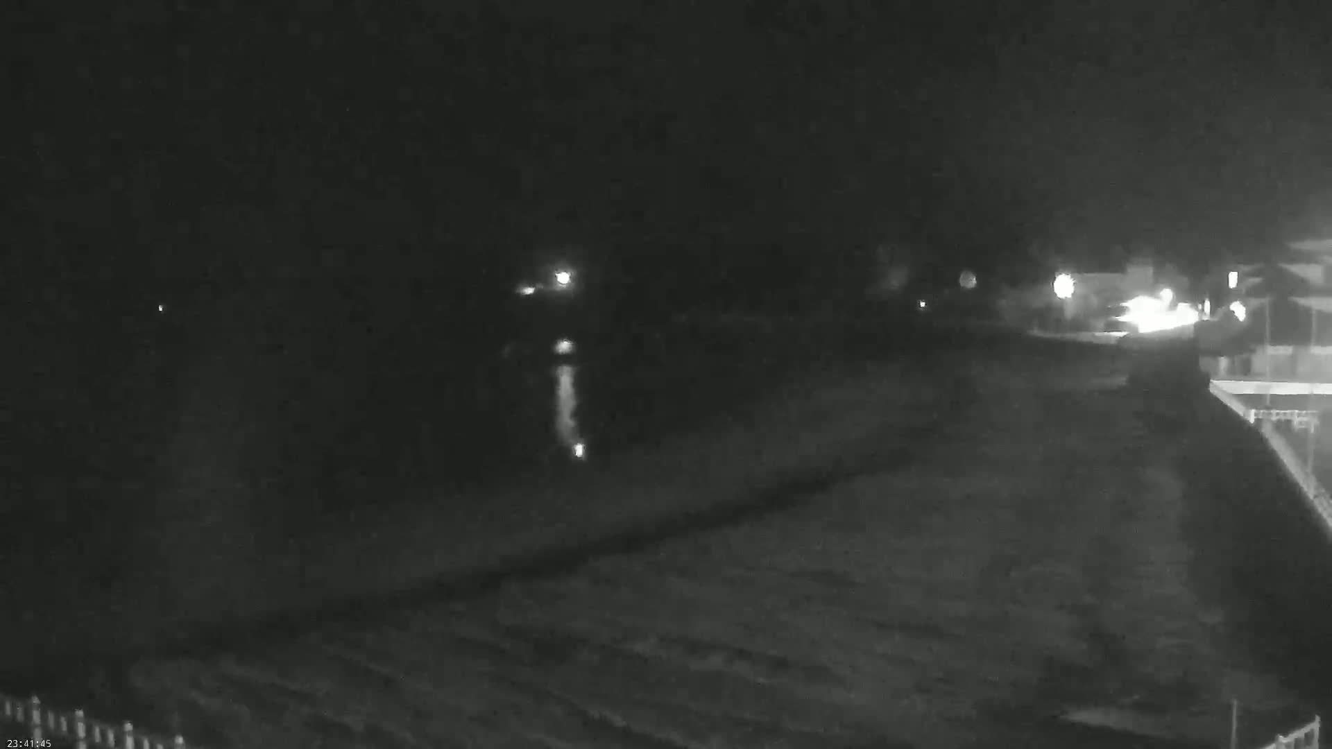 A nighttime, low-light view of a dark beach or shoreline with faint lights visible in the distance.