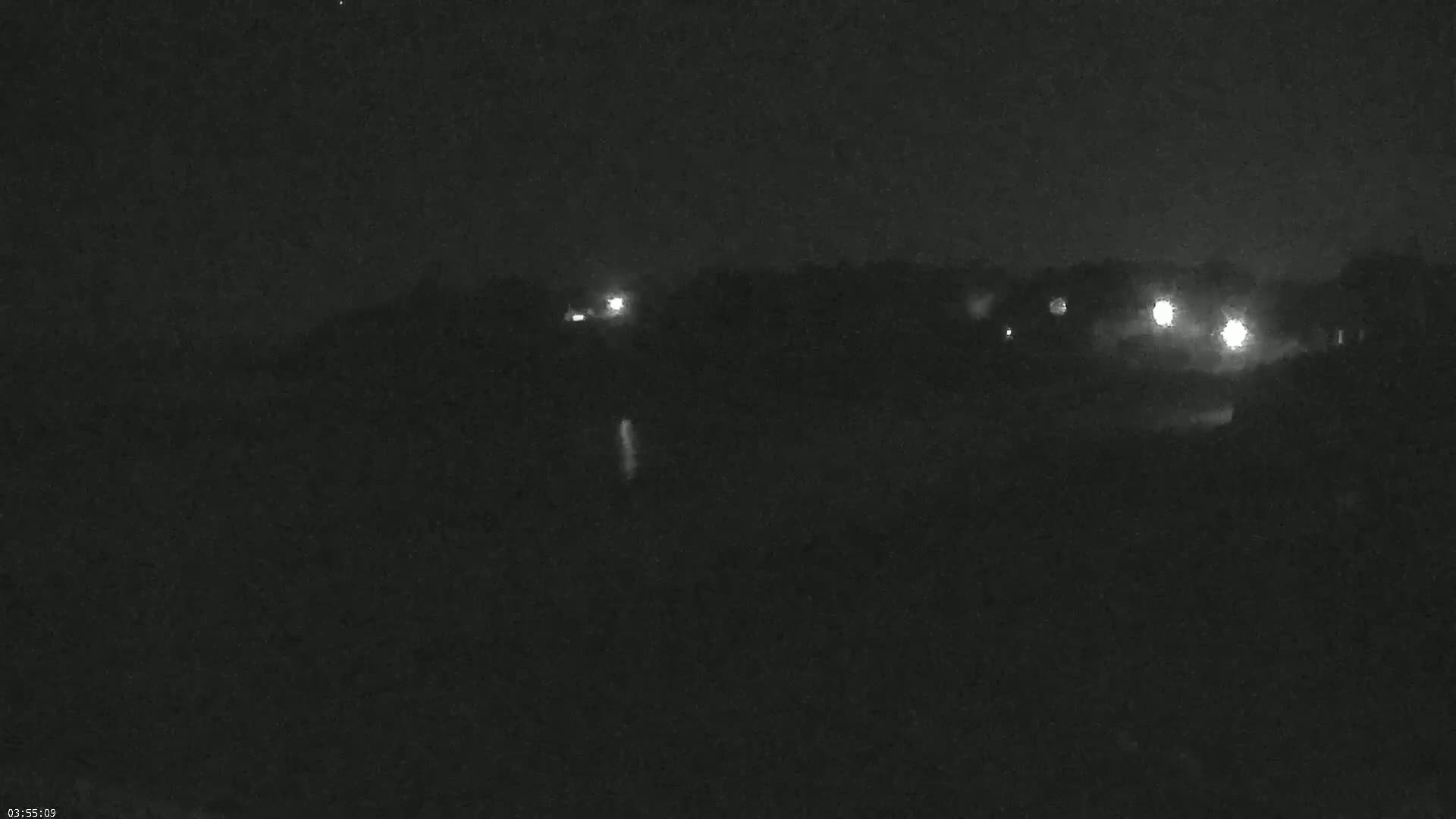 A nighttime, low-light image shows a dark, tree-lined landscape with several distant lights visible.