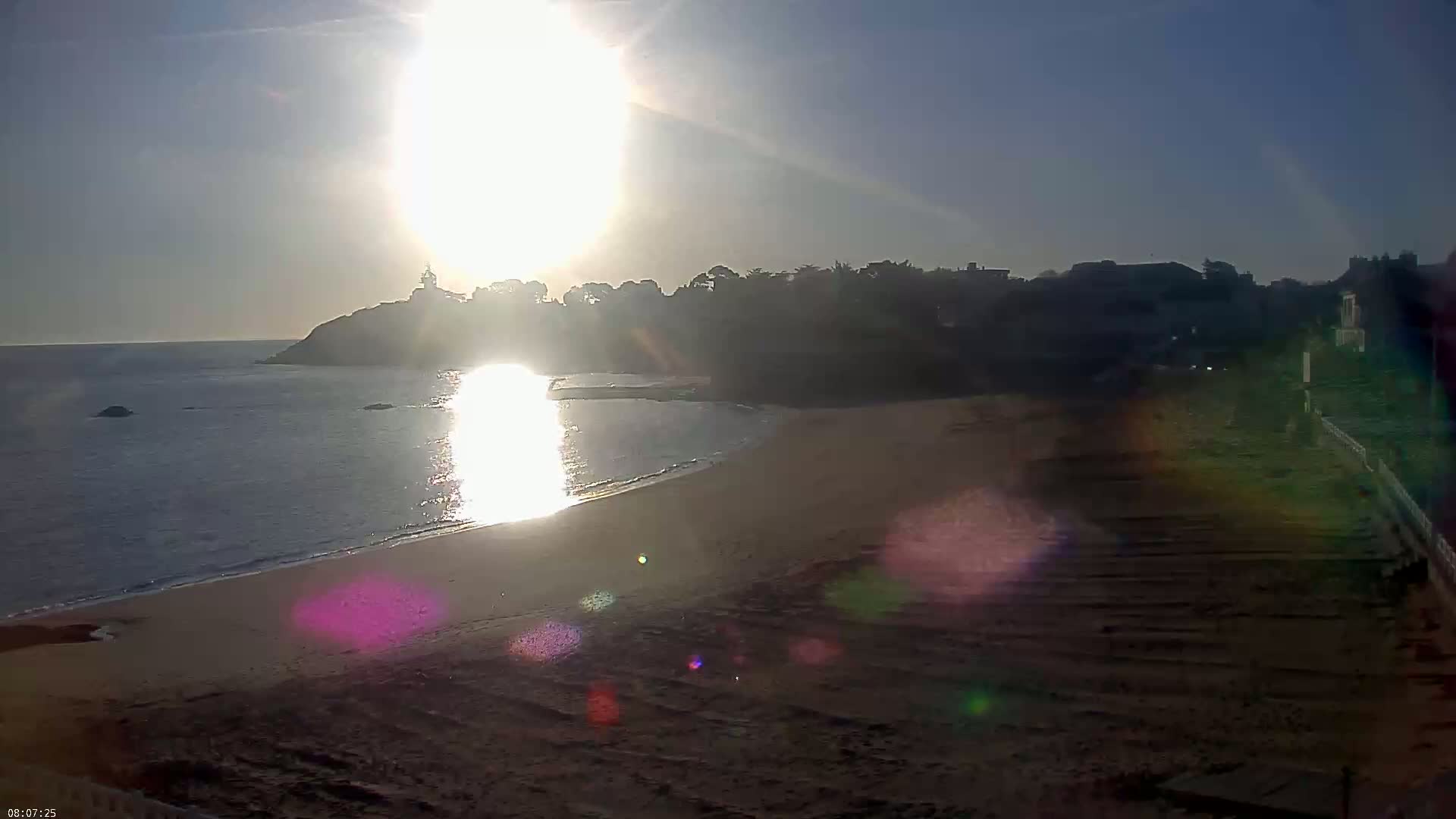 A sandy beach meets a calm ocean under a bright, sunny sky, with a distant coastline featuring buildings and a structure resembling a lighthouse.