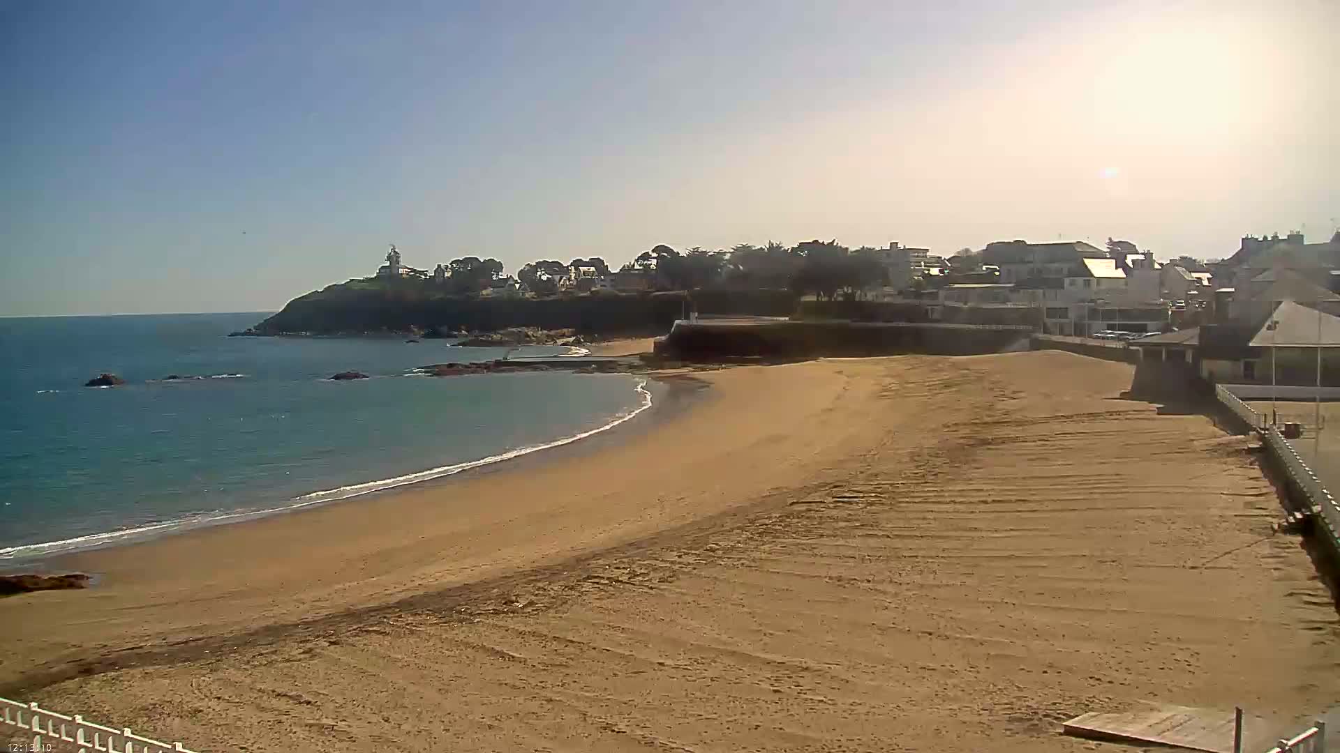 A sandy beach stretches out to a calm ocean under a clear, sunny sky, with a town and a small headland visible in the background.