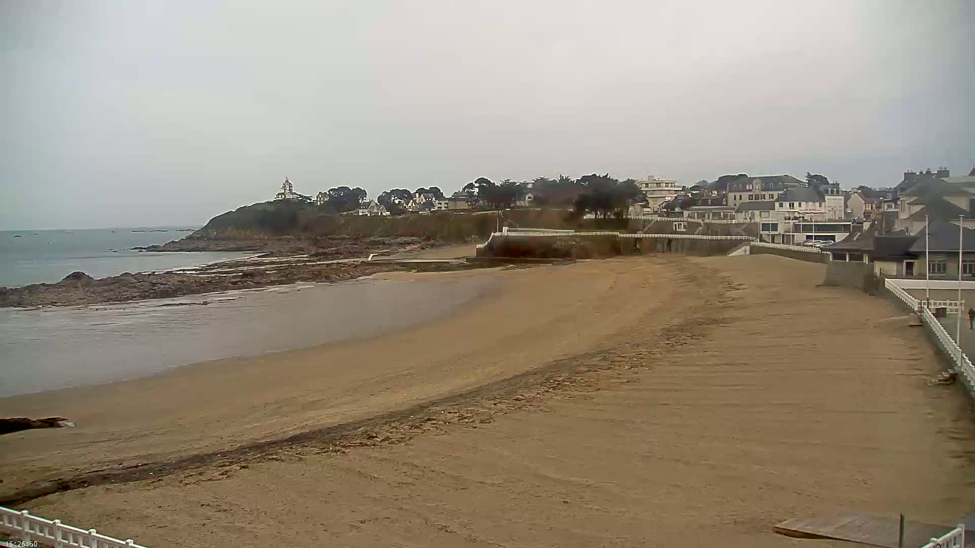 A sandy beach stretches towards a town situated on a cliff overlooking a calm sea under an overcast sky.