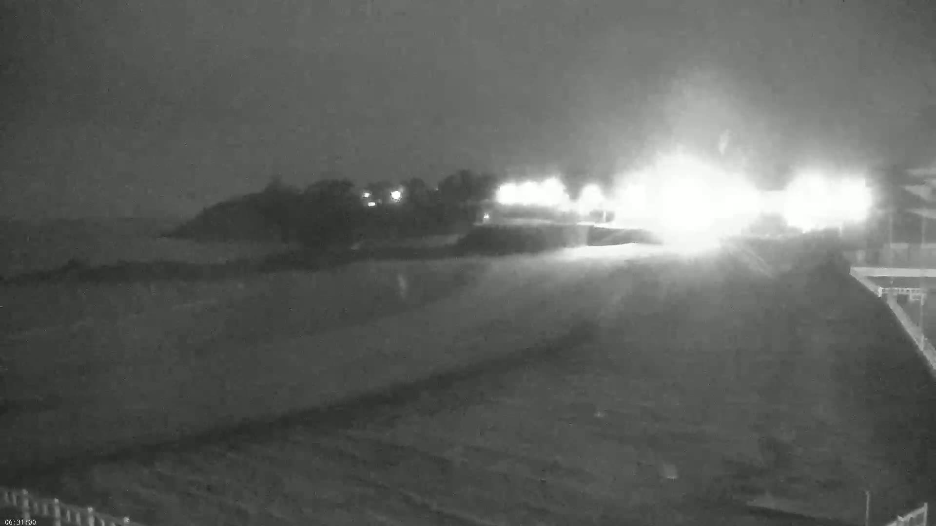 A nighttime, low-visibility view of a road with bright lights in the distance, possibly from oncoming traffic, and dark, indistinct land and water features nearby.