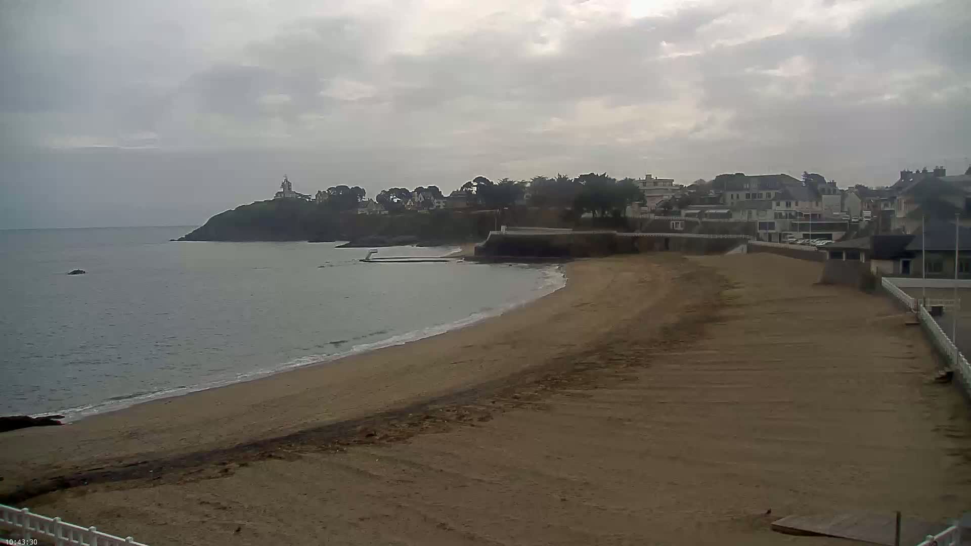 A sandy beach curves around a calm, grey sea towards a town nestled on a cliff, under an overcast sky.