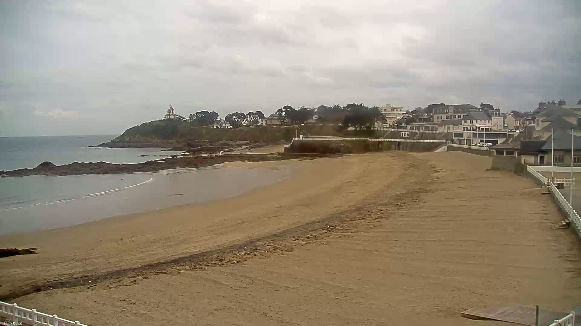 A sandy beach stretches towards a calm ocean, backed by a town and a hilltop structure under an overcast sky.