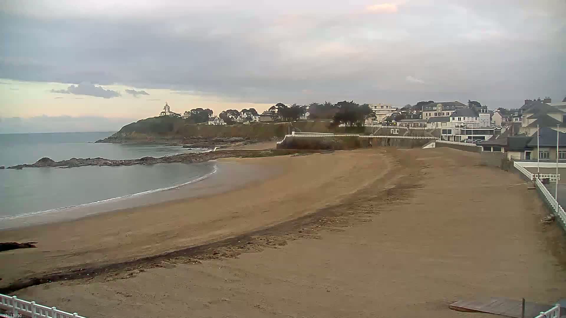A sandy beach stretches towards a coastal town under a cloudy sky, with calm ocean water lapping at the shore.