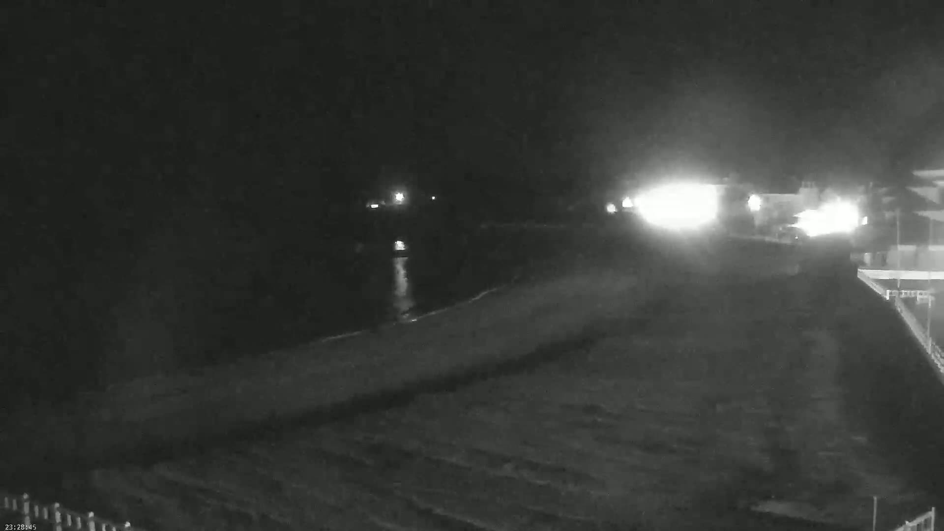 A nighttime, low-light view shows a dark body of water reflecting lights from a roadway and buildings beyond a shoreline.