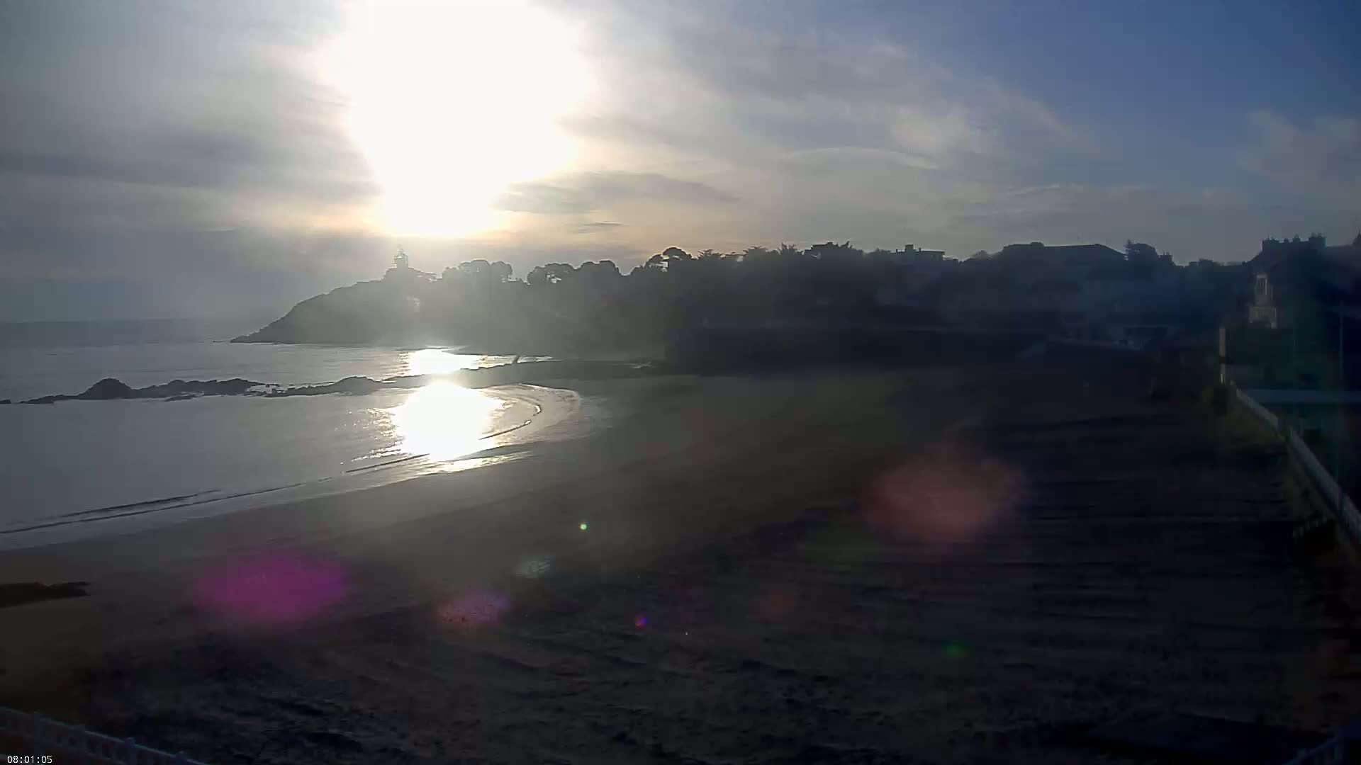 A sandy beach and calm ocean are seen at sunrise, with a small town and a lighthouse visible in the distance.