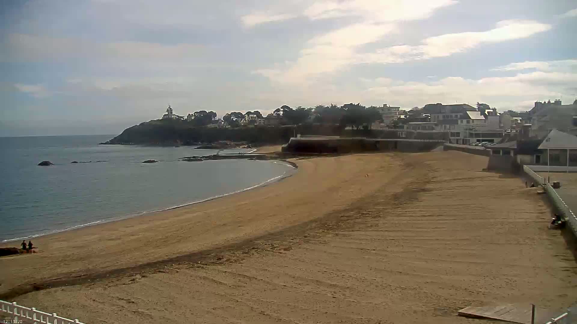 A sandy beach curves gently around a calm ocean, with a small town and a lighthouse visible on a nearby hill under a partly cloudy sky.