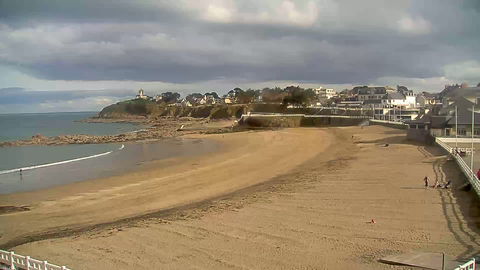 A sandy beach stretches out to a calm ocean under a cloudy sky, with a small town and a lighthouse visible in the distance.