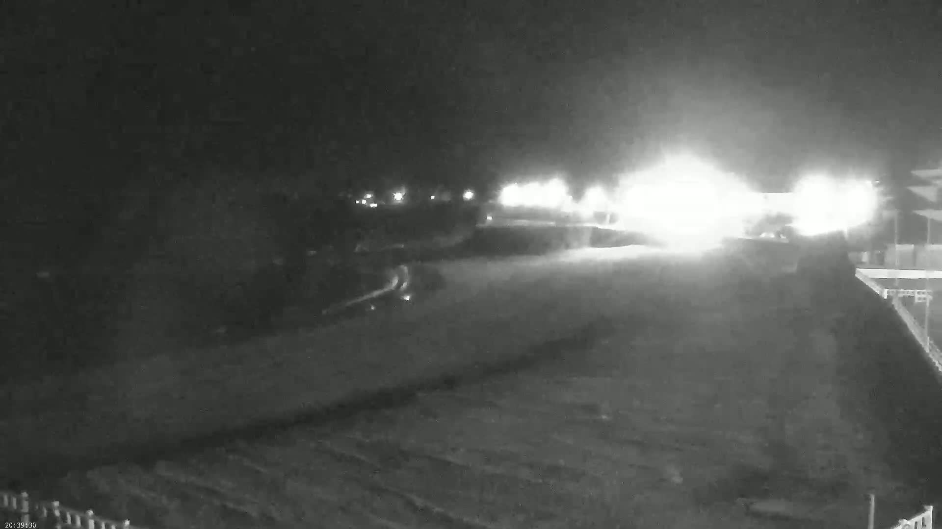 A nighttime, low-light view shows a paved area with bright lights in the distance, possibly a road or parking lot, next to a dark, unpaved area.