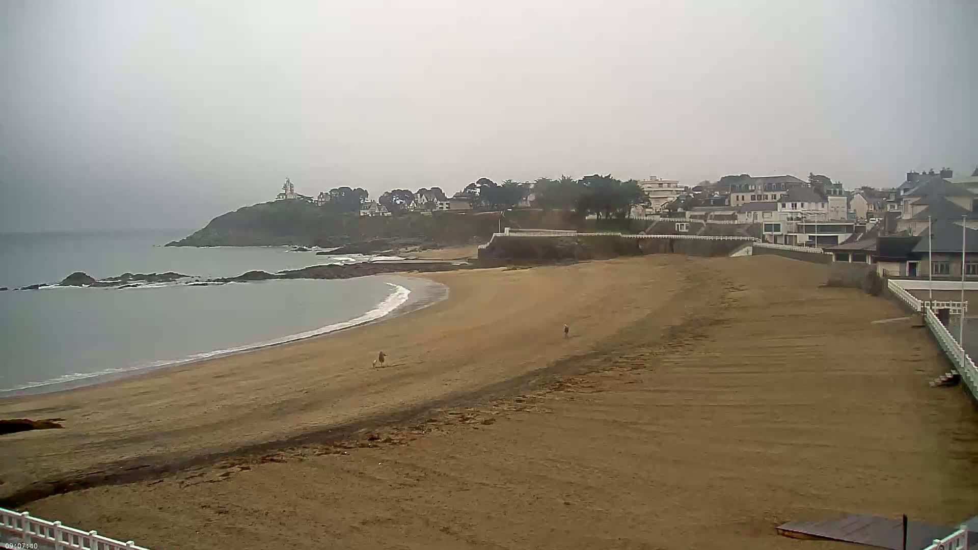 A sandy beach curves around a calm, grey ocean under an overcast sky, with a small town and a lighthouse visible on a distant hill.