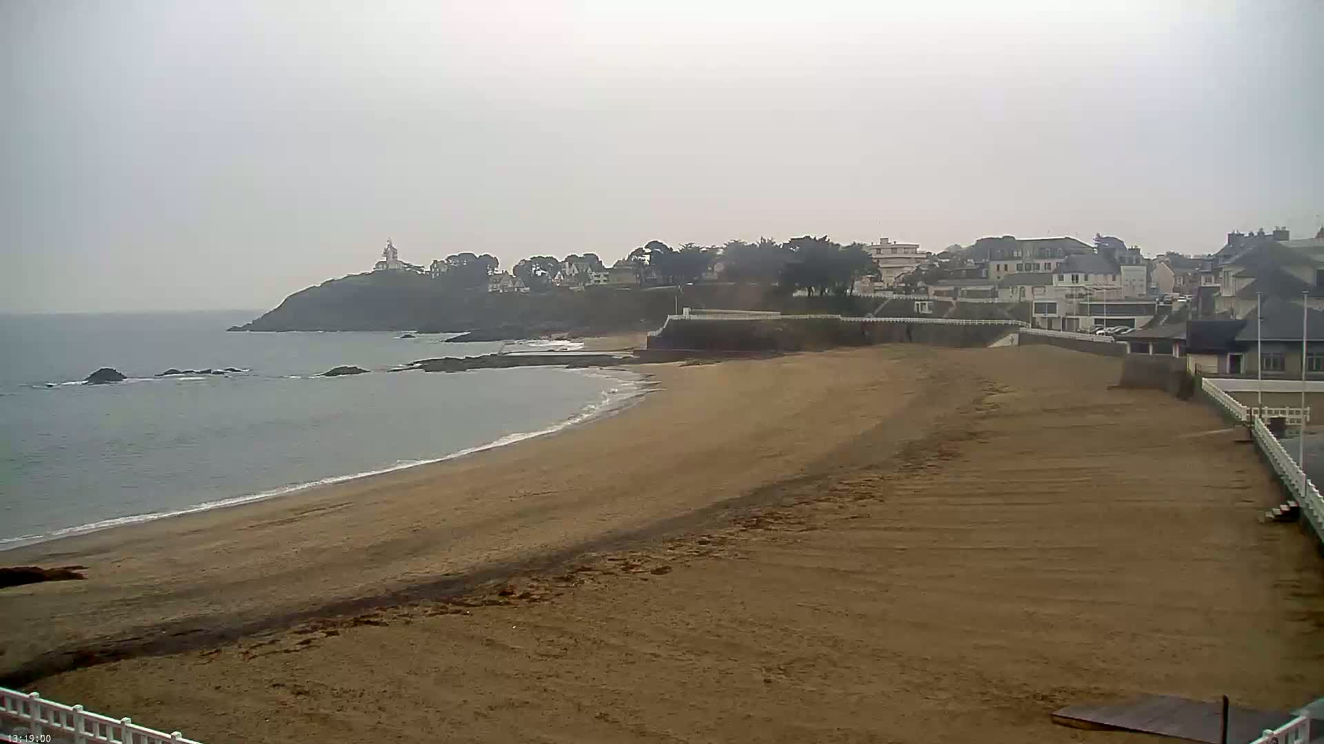 A sandy beach stretches towards a calm ocean under an overcast sky, with a small town and a lighthouse visible in the distance on a cliff.