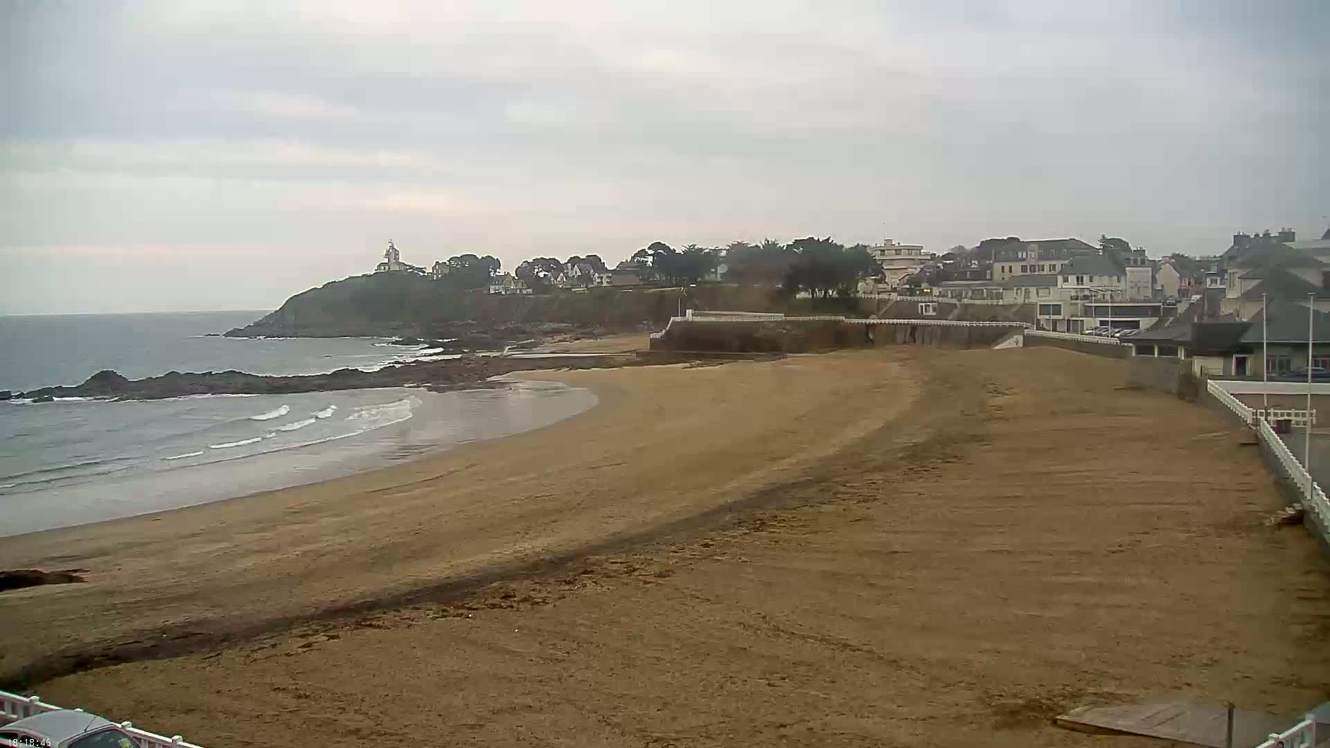 A sandy beach curves gently alongside a rocky coastline, featuring a small town and a distant lighthouse under an overcast sky.