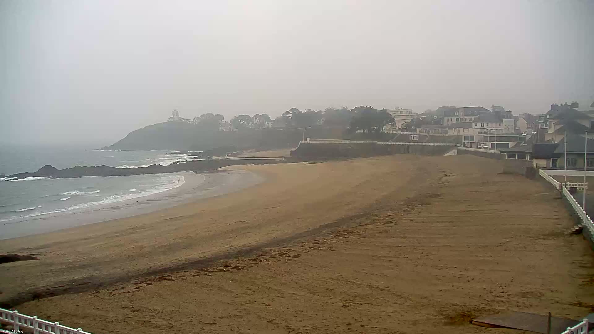 A sandy beach stretches out to a calm, foggy ocean, bordered by a low cliff with houses and a lighthouse in the distance.
