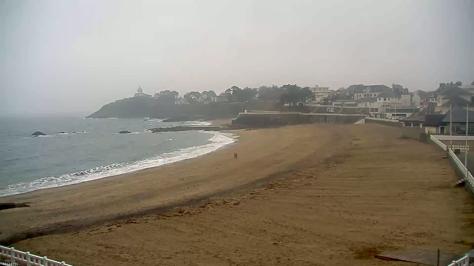A wide sandy beach stretches towards a hazy coastal town under a foggy sky, with small waves lapping the shore.