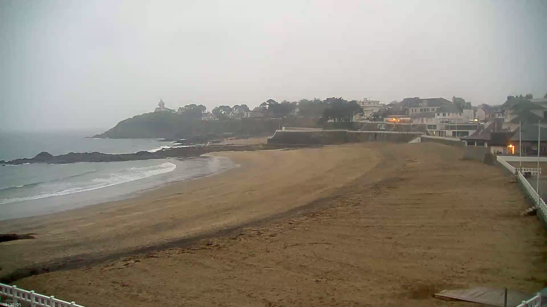 A sandy beach with gentle waves stretches towards a coastline featuring buildings and a lighthouse under an overcast sky.