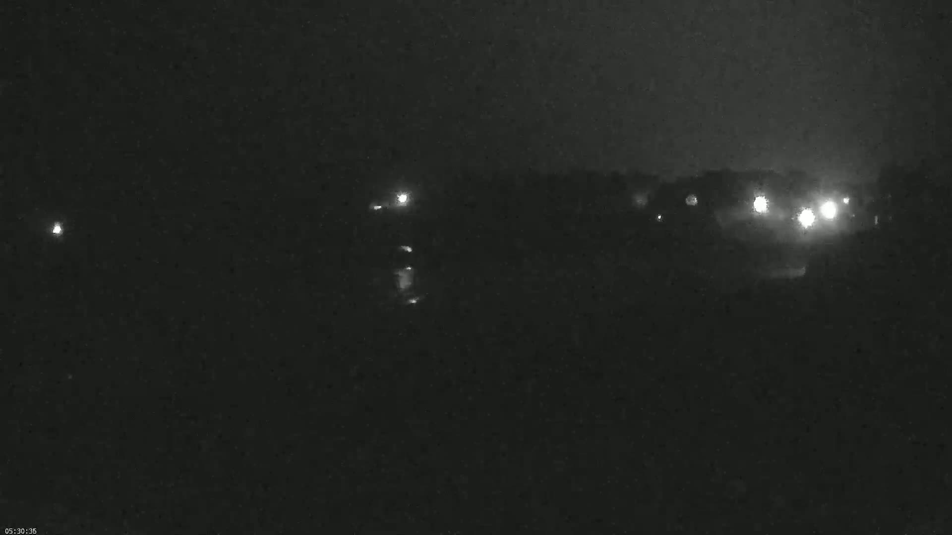 A nighttime, dark scene shows a few distant lights, possibly from vehicles or buildings, under a cloudy sky.