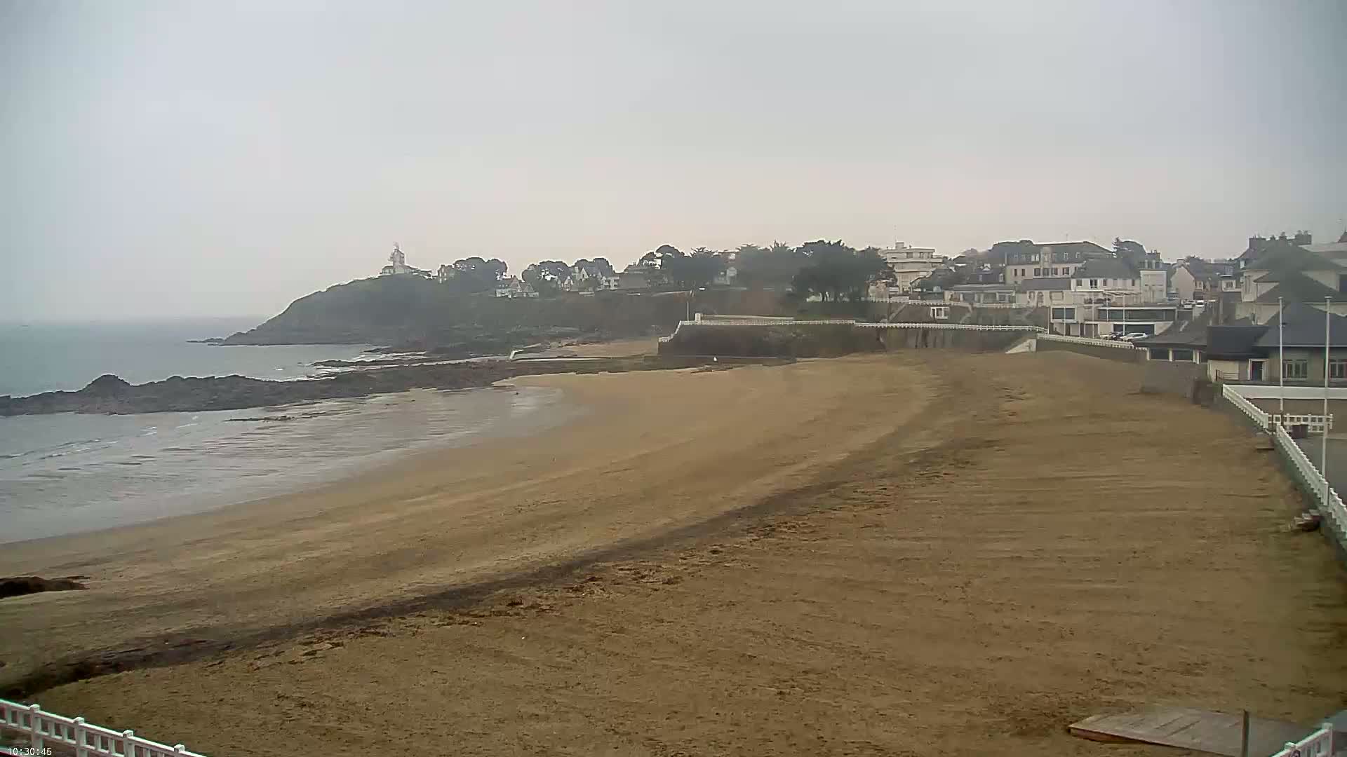 A sandy beach stretches out to a calm sea under an overcast sky, with a coastal town and a lighthouse visible on a nearby hill.