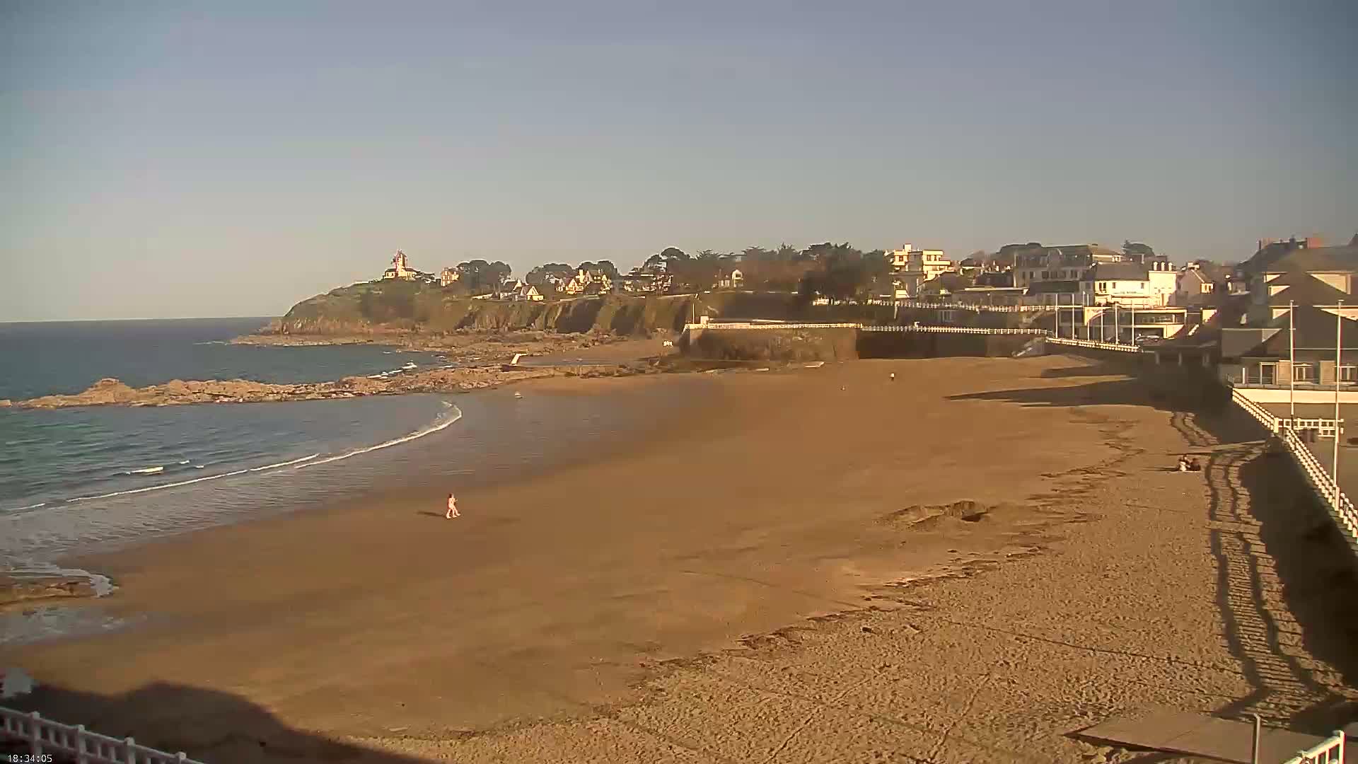 A sandy beach stretches out to a calm ocean under a clear sky, with a small town and a cliff visible in the background.