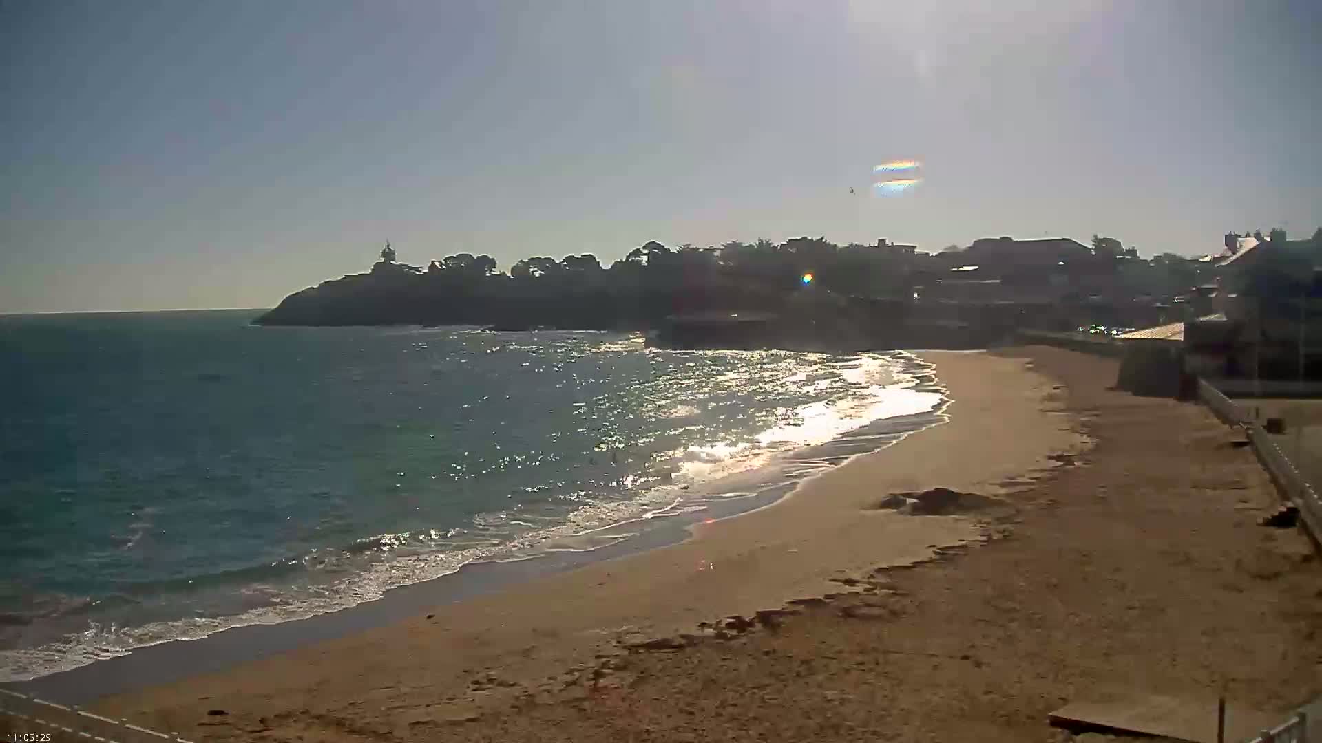 A sandy beach with small waves laps the shore under a bright, sunny sky, with a small town and a cliff with a lighthouse visible in the distance.
