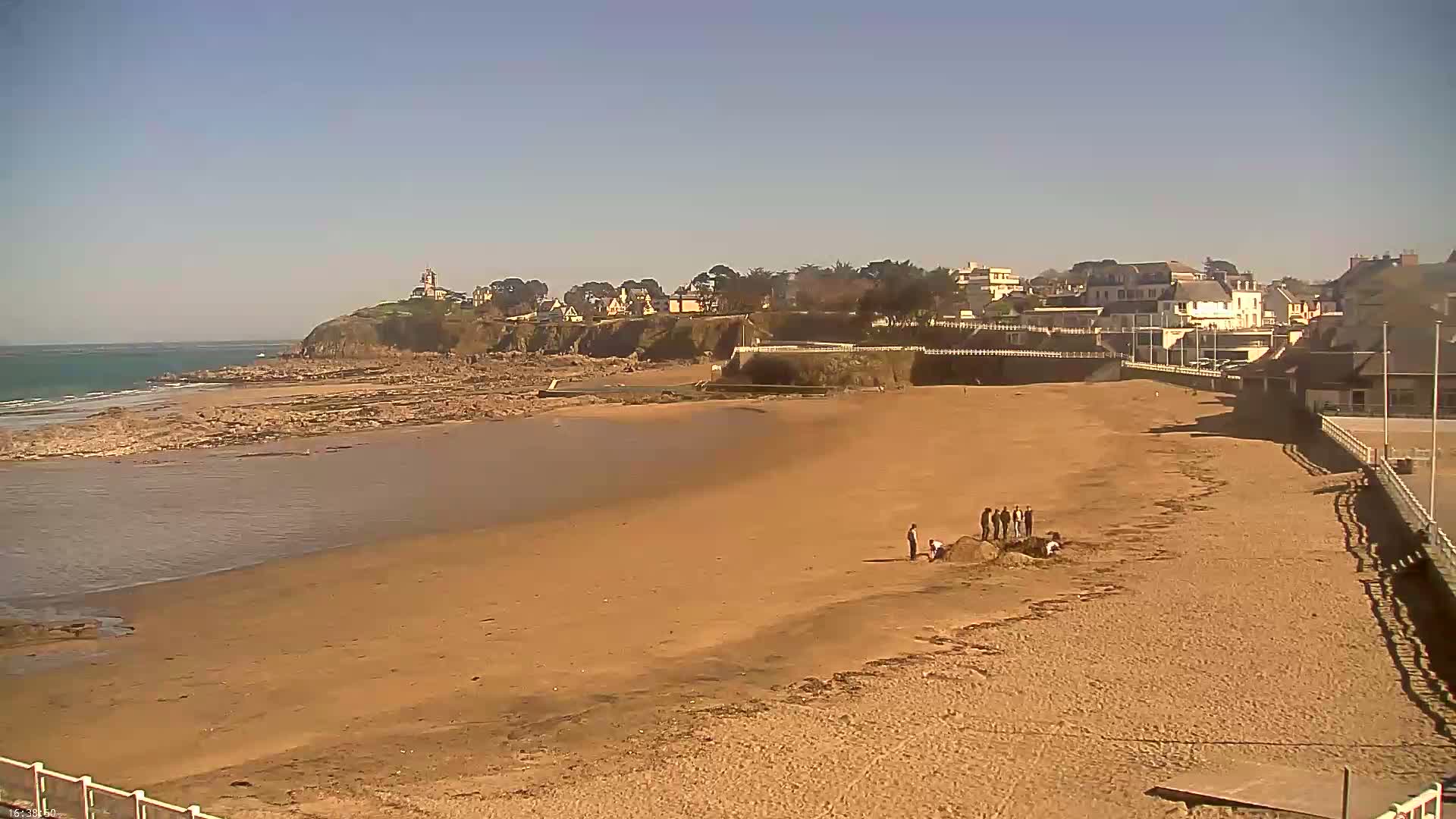 A sandy beach stretches out to a calm ocean under a clear, sunny sky, with a small group of people near the water's edge and buildings perched on a cliff in the background.