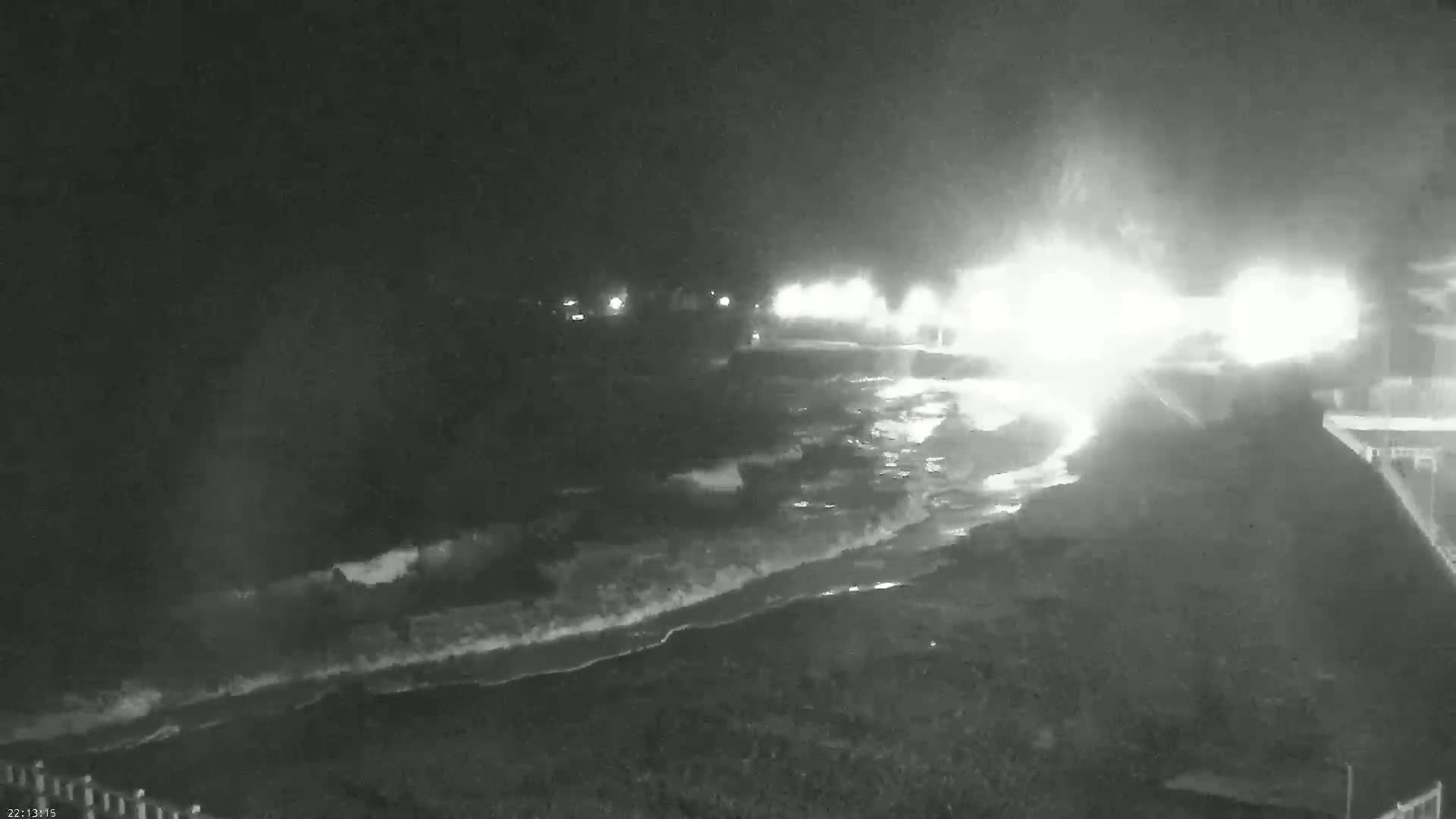 A nighttime, low-light beach scene shows waves lapping the shore under a dark, possibly stormy sky, illuminated by bright lights in the distance.
