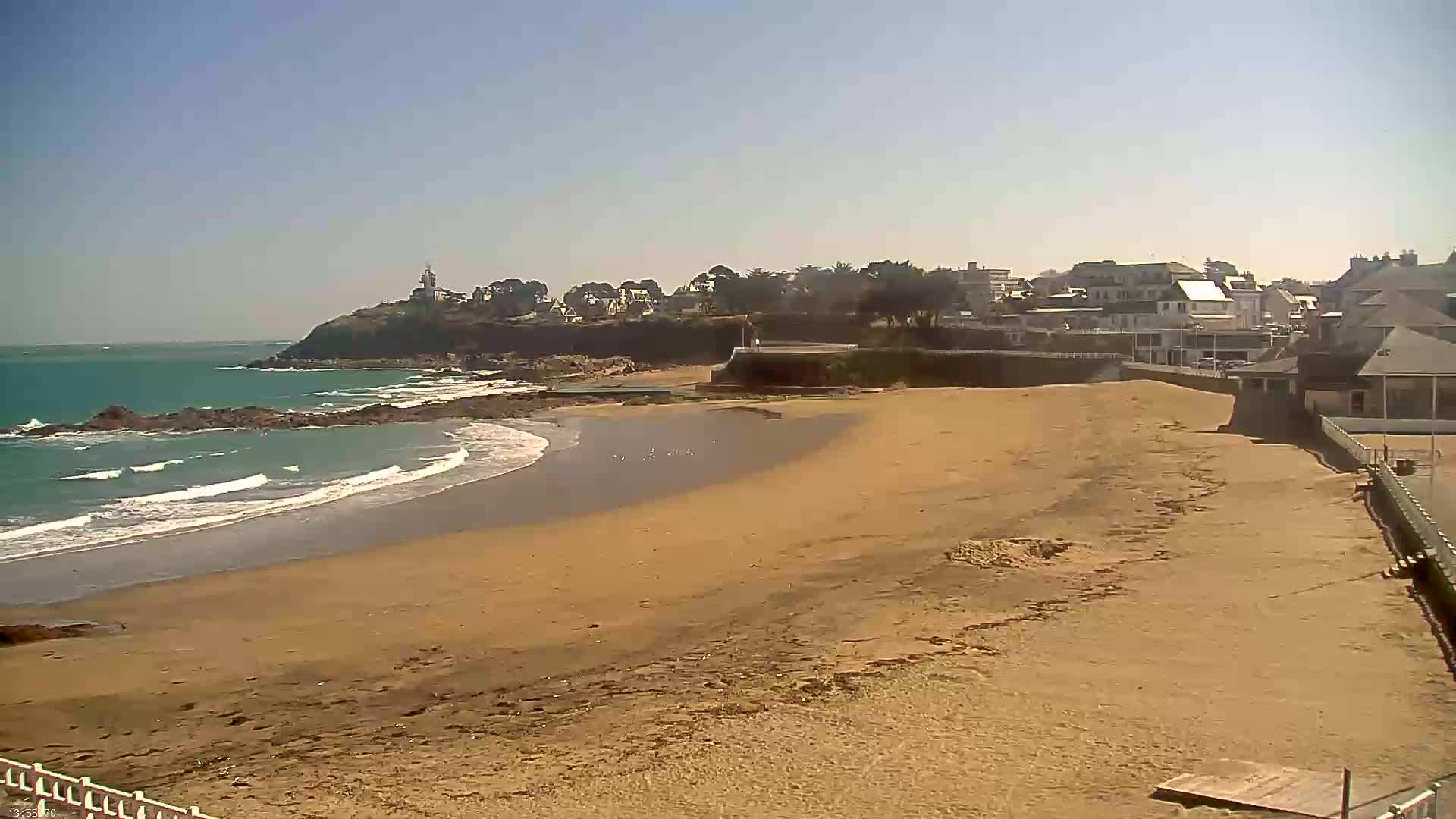 A sandy beach stretches from a calm, teal ocean toward a town nestled on a rocky cliff, under a clear, sunny sky.