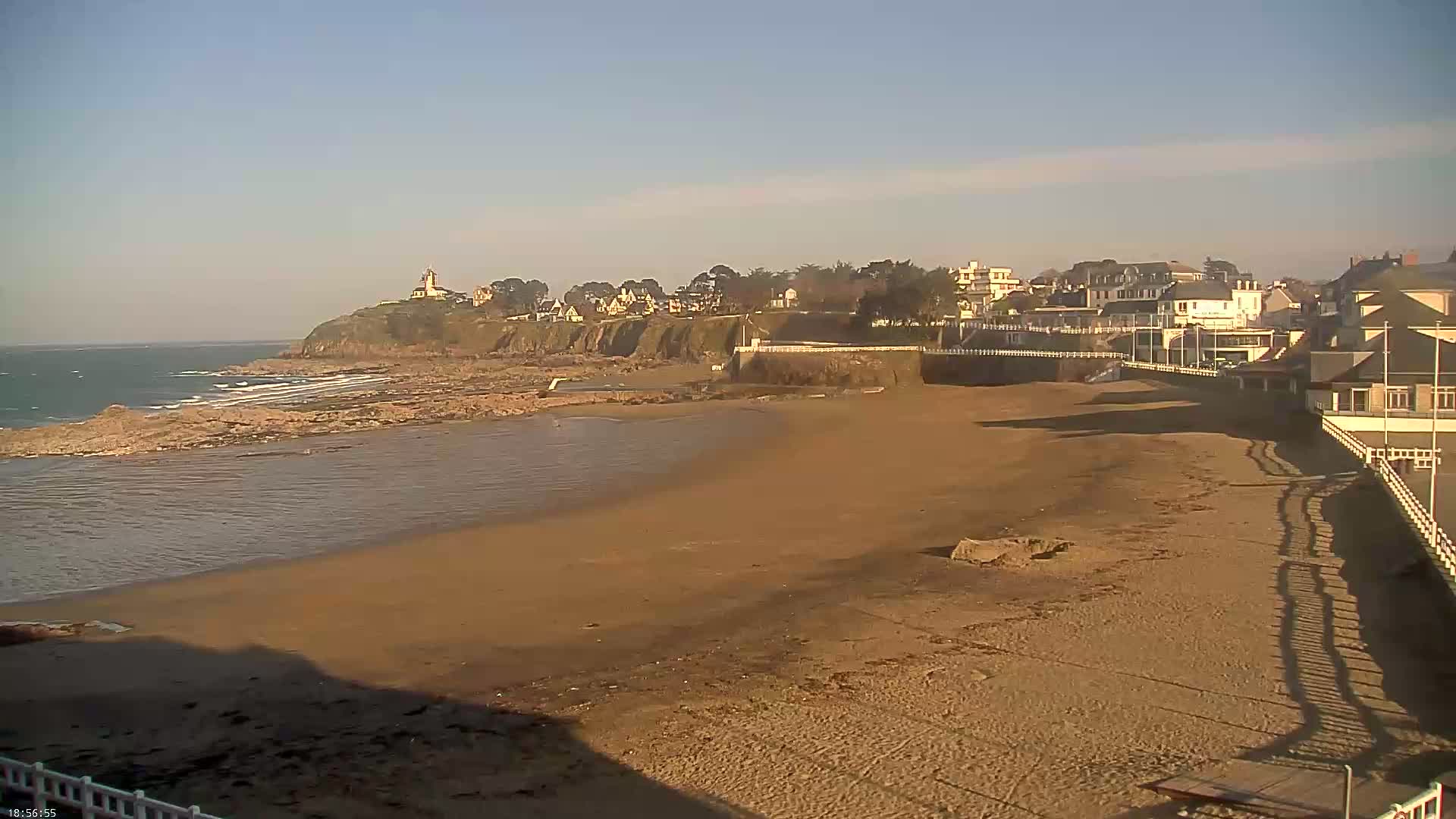 A sandy beach stretches out to a calm ocean under a clear, bright sky, with a line of buildings and a rocky headland visible in the distance.