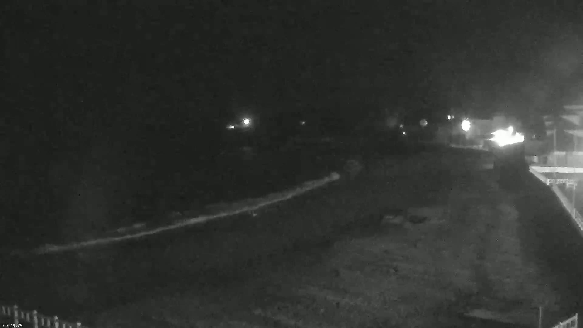 A nighttime, low-light outdoor scene shows a long, bright streak across a dark, possibly snowy or icy ground, with distant lights visible in the background.