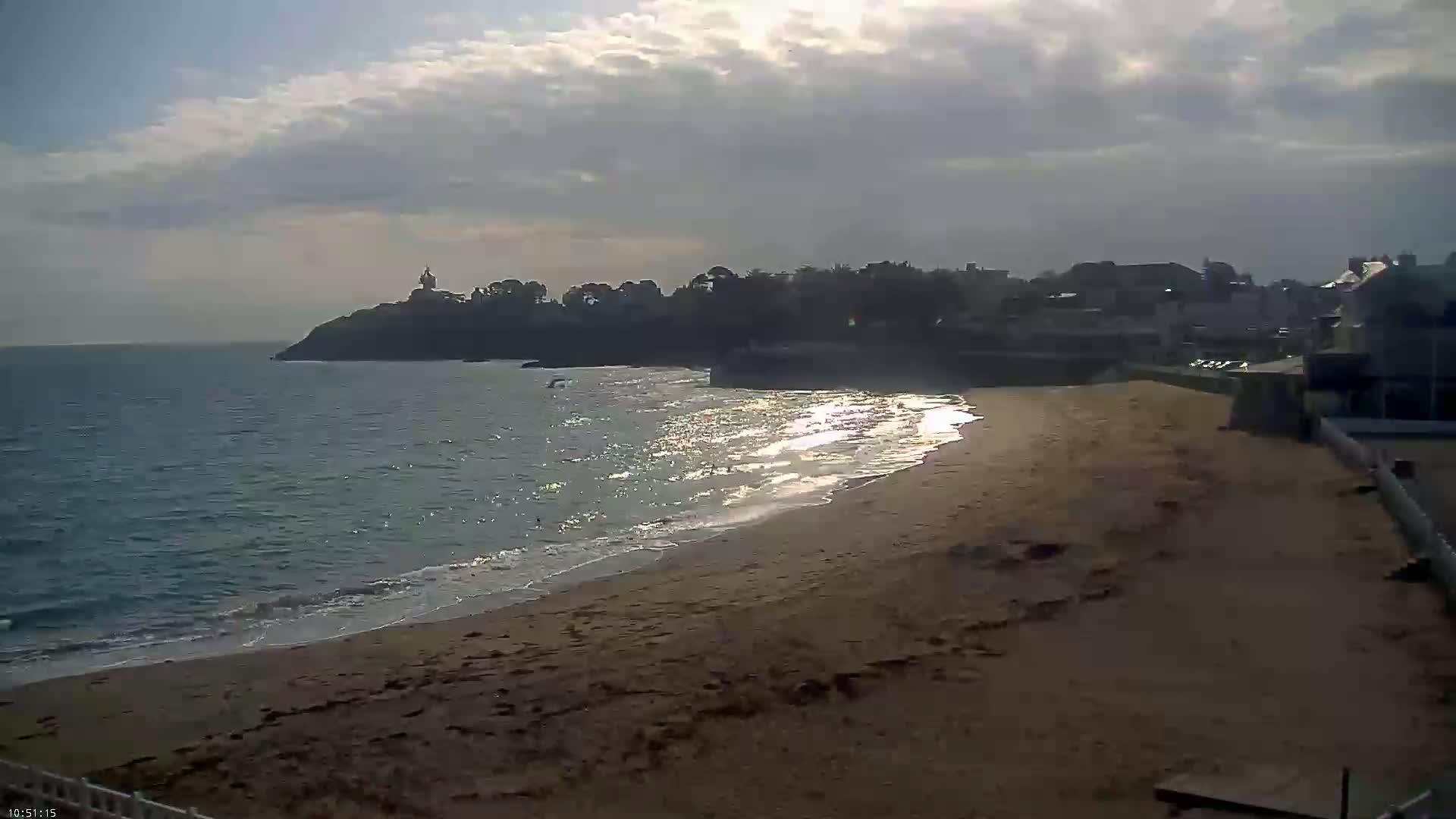 A sandy beach with gentle waves laps the shore under a partly cloudy sky, with a town and a distant lighthouse visible on a nearby headland.