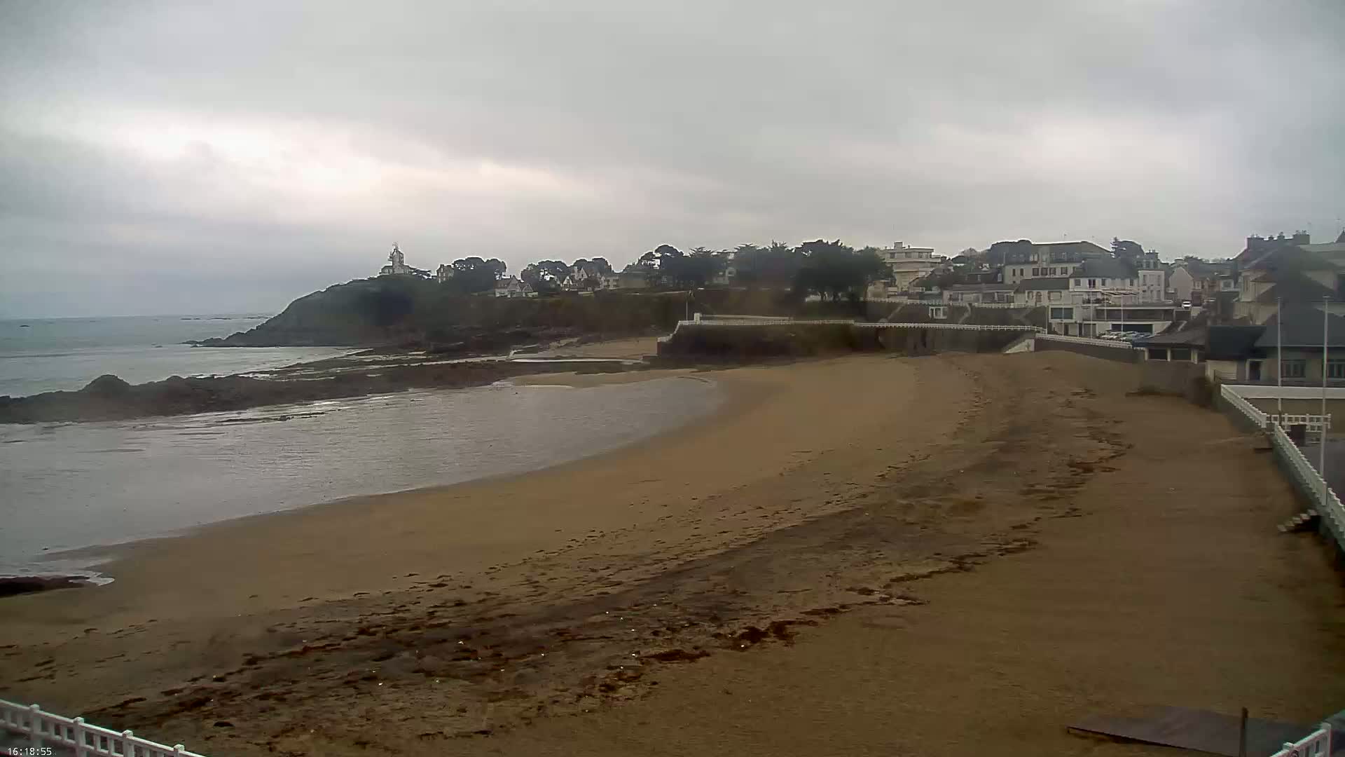 A sandy beach curves around a calm bay, under an overcast sky, with buildings and a small lighthouse visible on a hill in the distance.