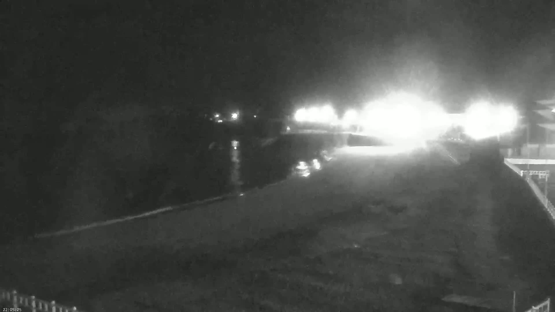 A nighttime, low-light view shows a body of water reflecting bright lights from a building across the water, possibly a road or parking area.