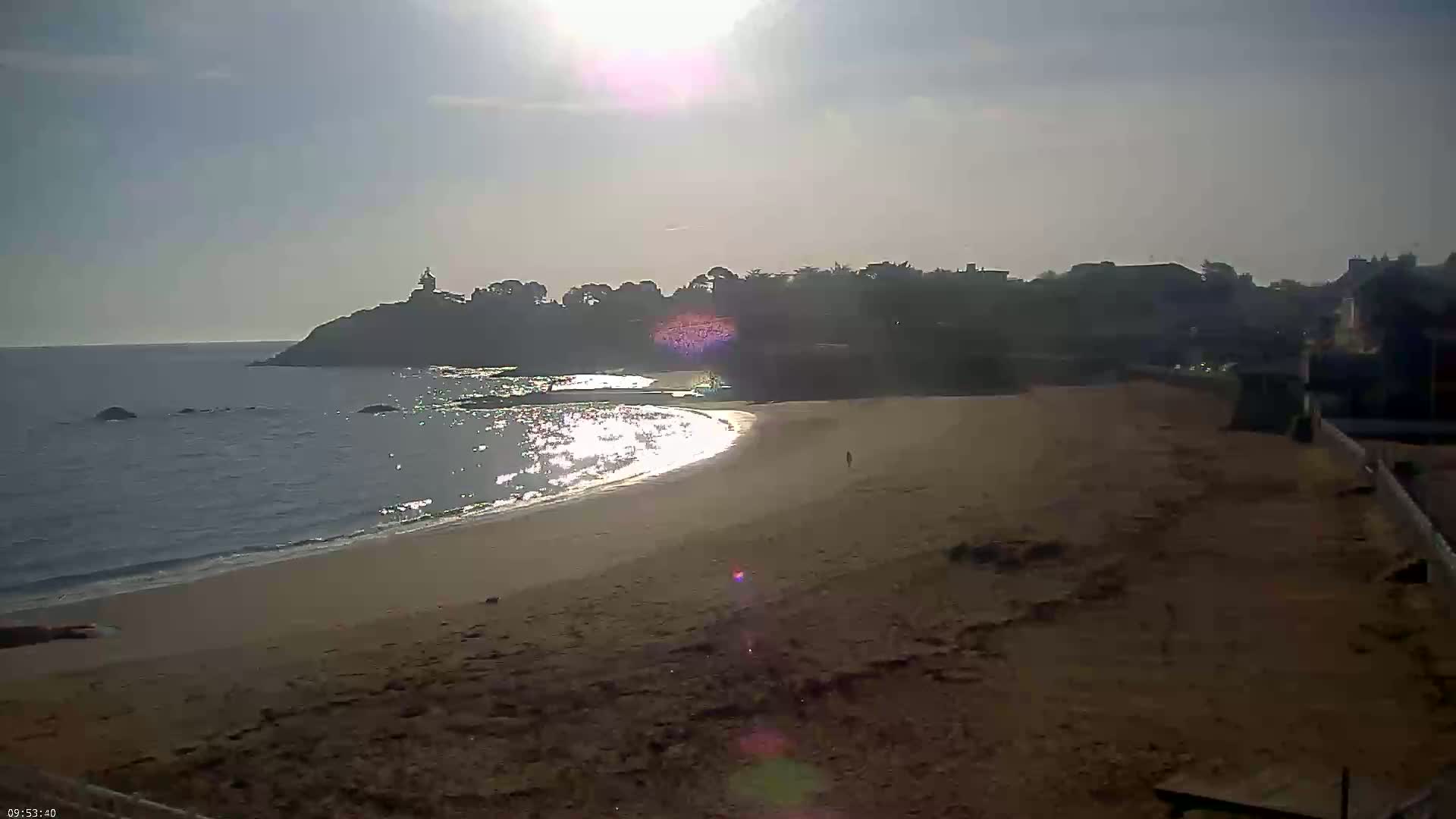 A sandy beach curves gently around a calm ocean under a bright, sunny sky, with a distant headland and buildings visible in the background.