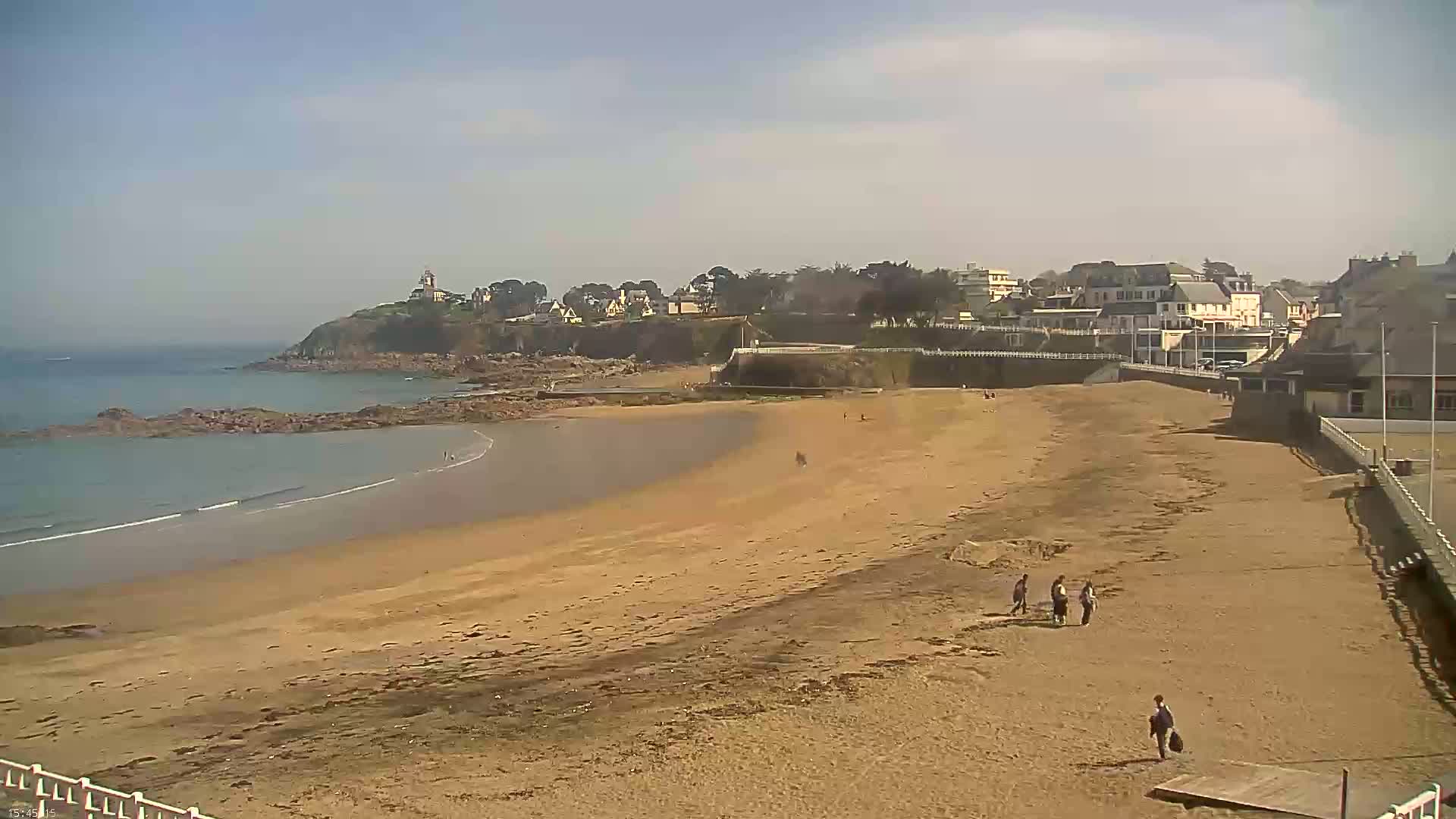 A sandy beach stretches out to a calm ocean, with a small town and a lighthouse visible on a hill in the background under a mostly clear sky.