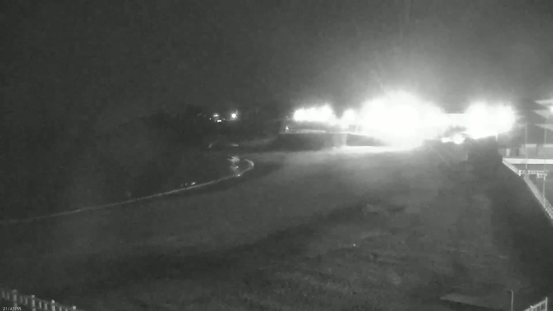 A nighttime, low-light view shows a paved area brightly lit in the distance, with darker, less-defined areas surrounding it.