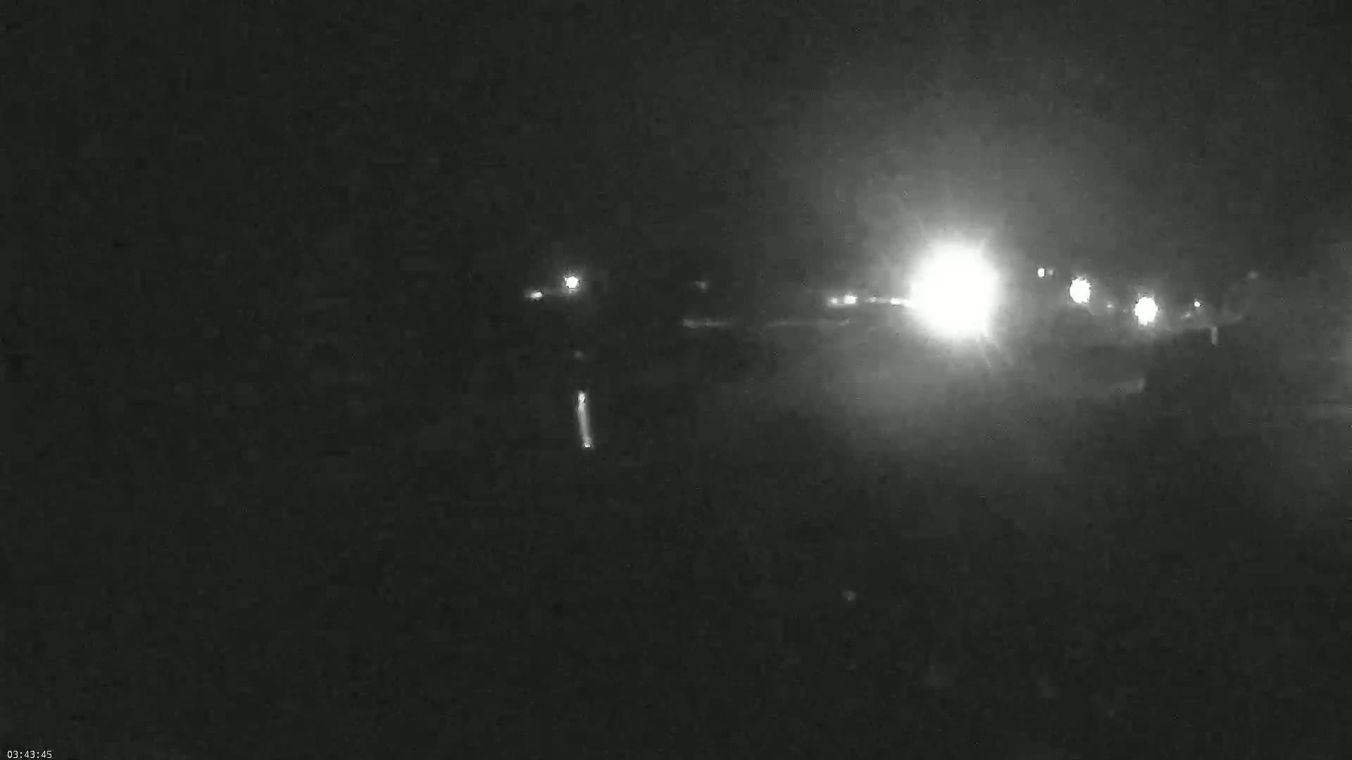 At night, bright headlights illuminate a dark, possibly foggy road with indistinct vehicles and lights in the distance.