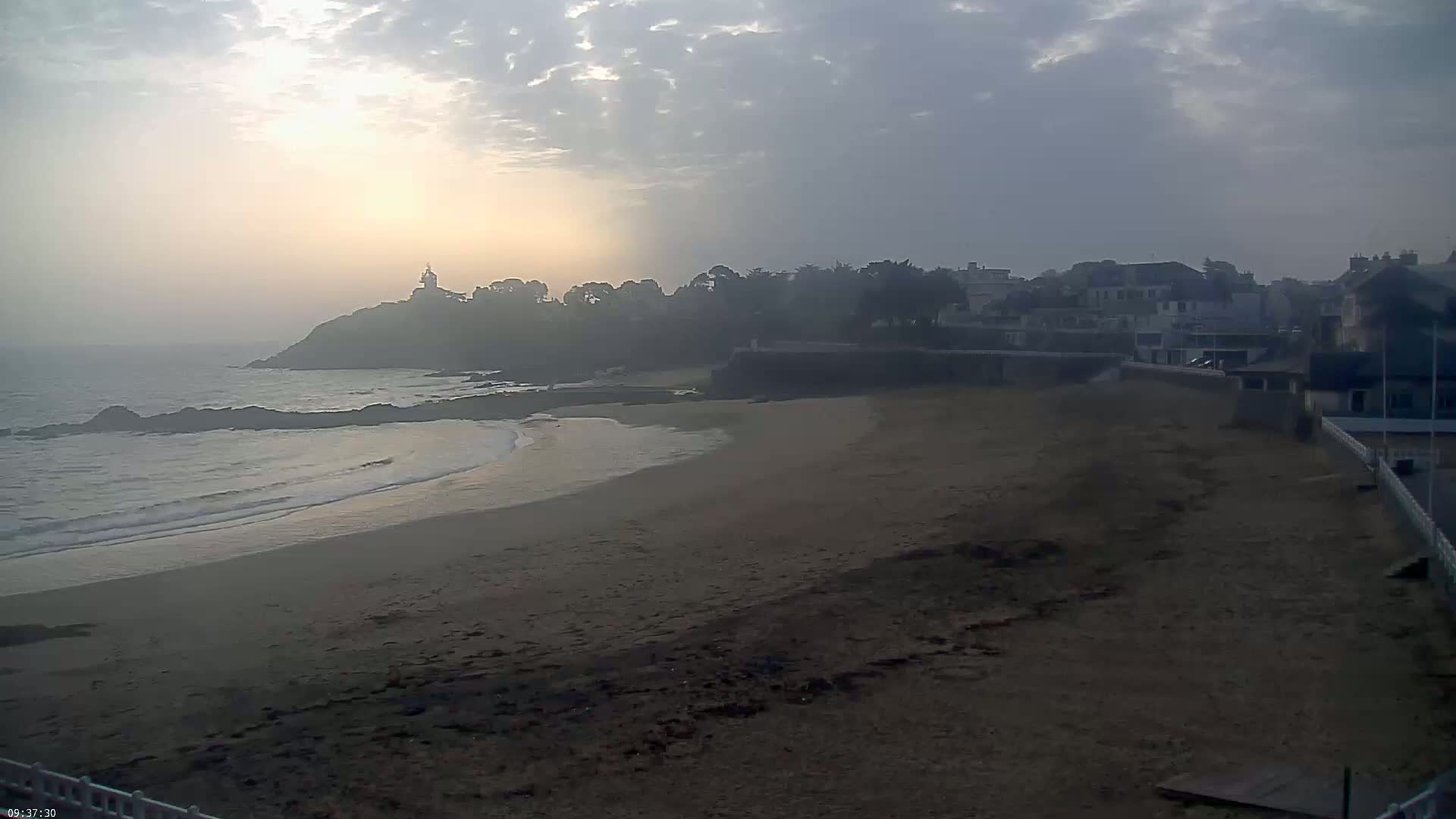 A hazy sunrise illuminates a sandy beach and a small town nestled beside a rocky coastline.