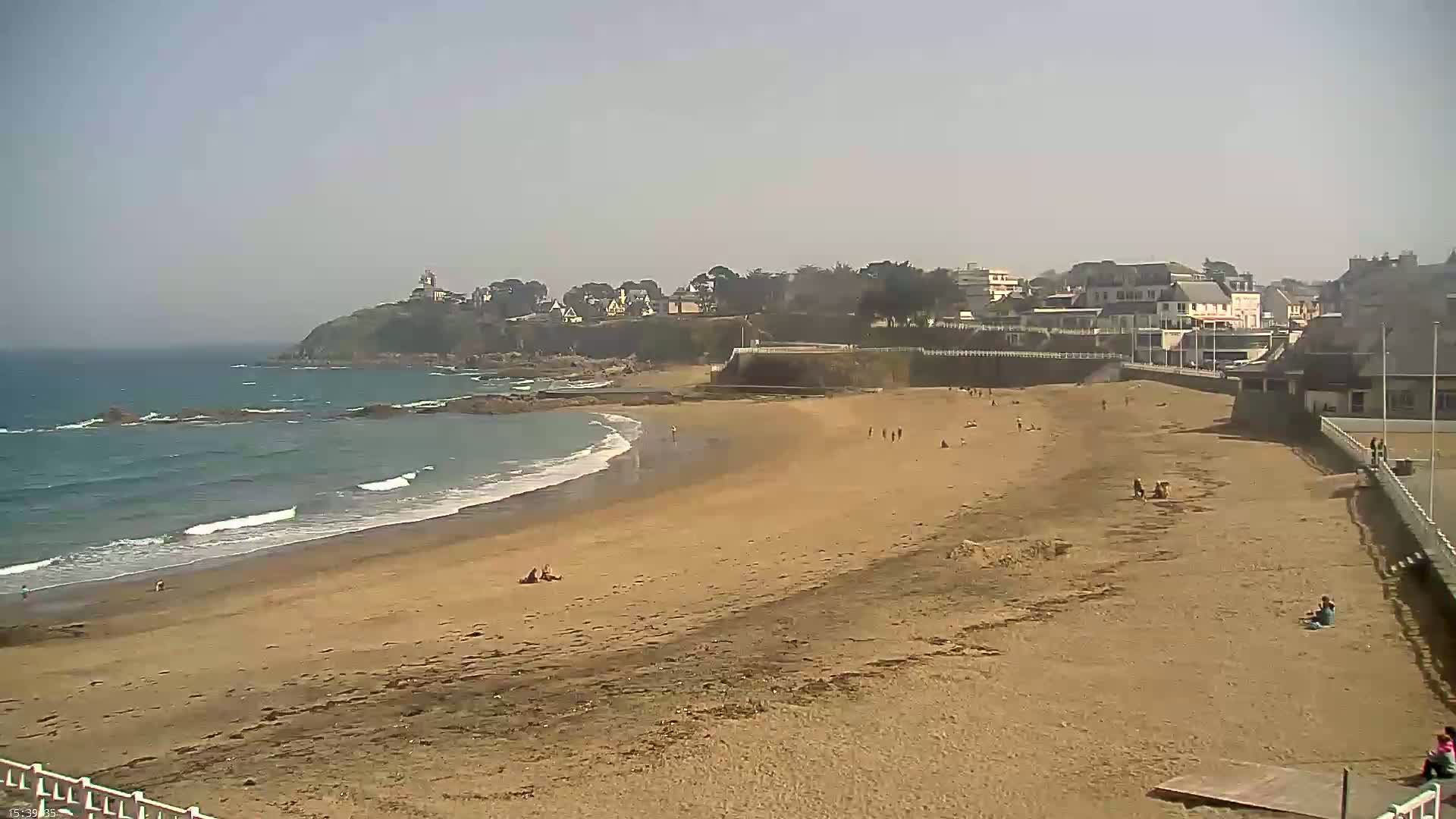 A sandy beach stretches out to a calm ocean with small waves under a clear, bright sky, backed by a town and a rocky headland.